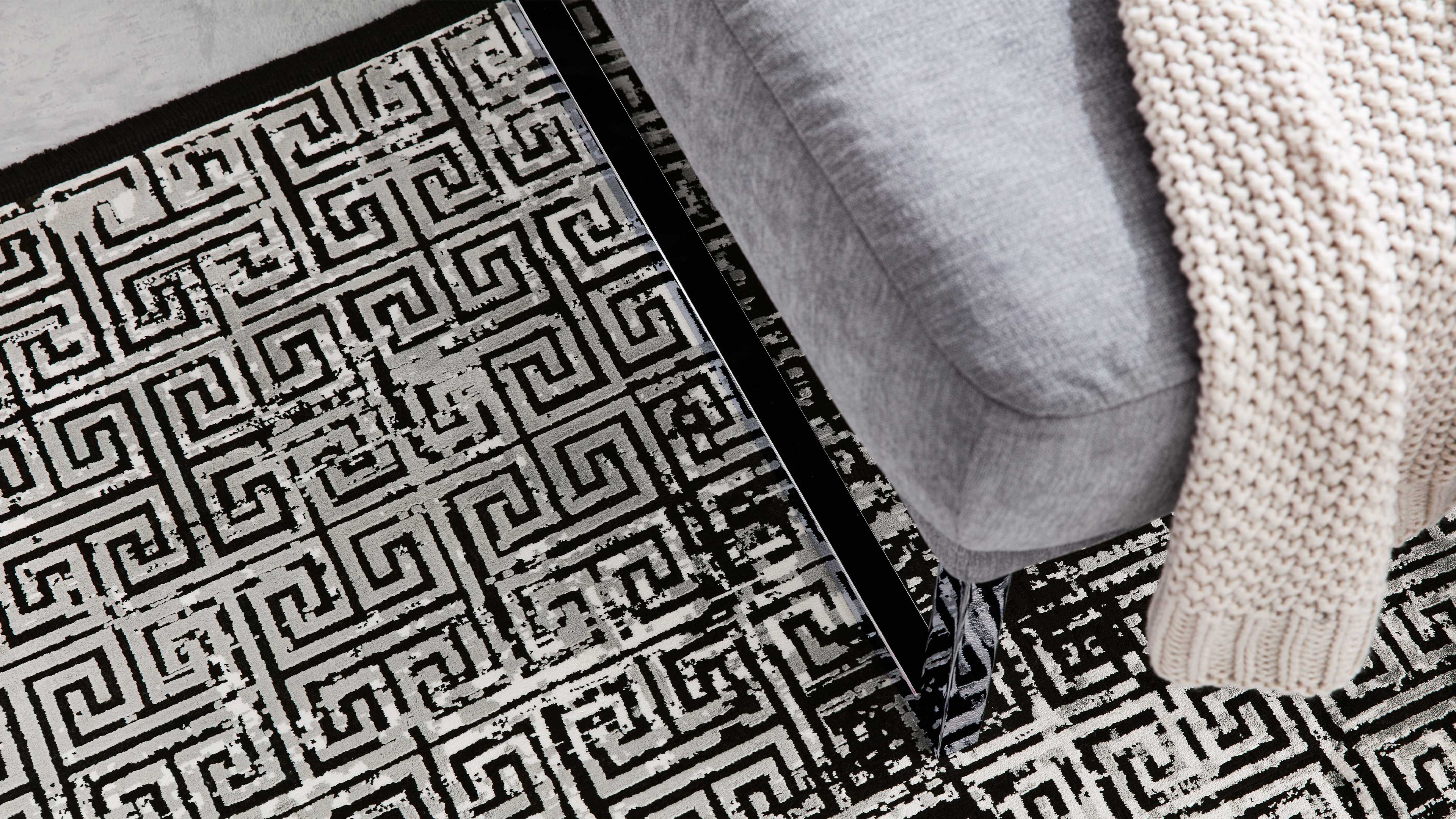 Grau bezogene Sofa-Ecke mit schwarzem Metallfuß, beigefarbener Grobstrickdecke und schwarz-weißem Teppich mit labyrinthischem Muster.