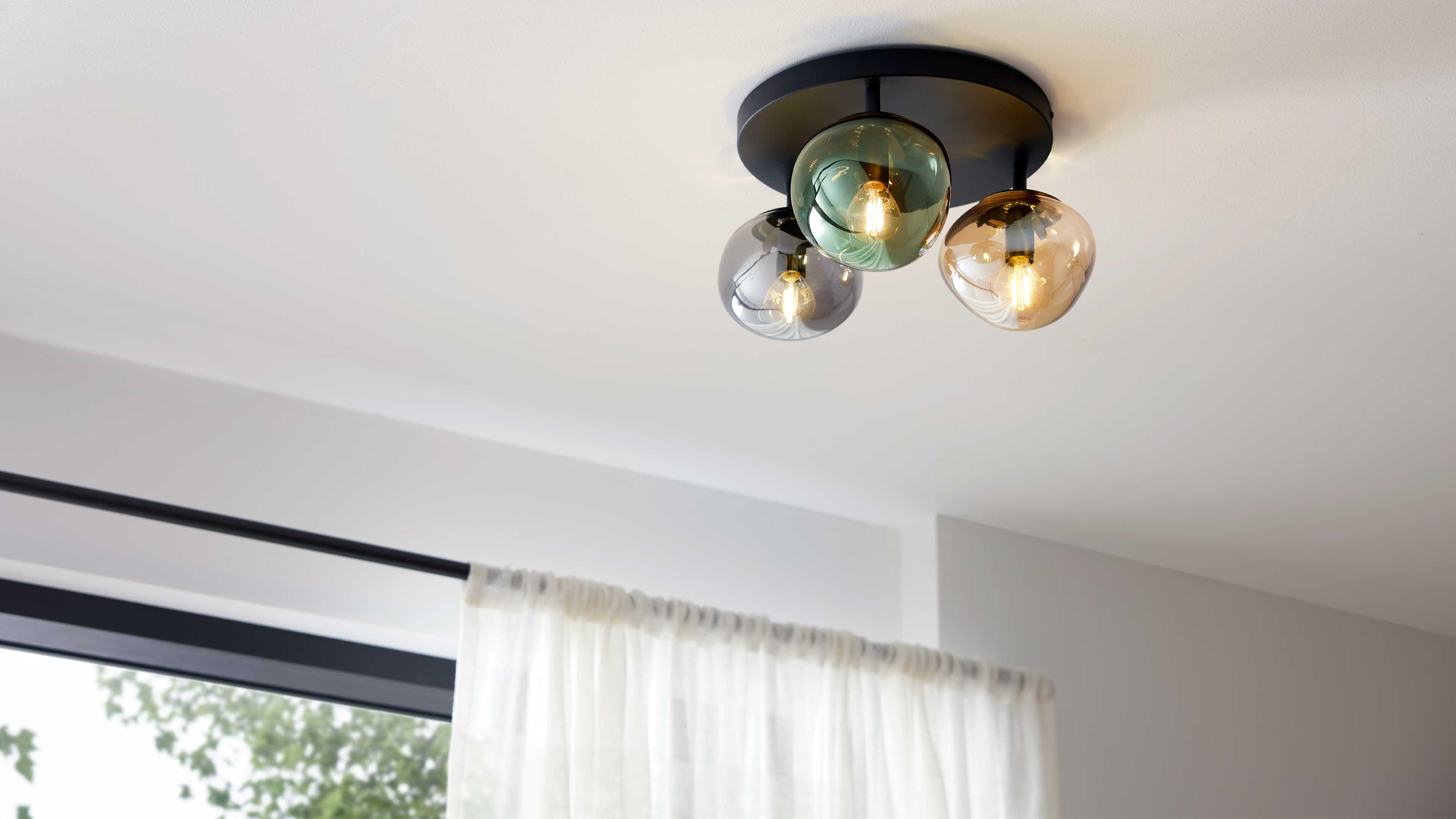 Deckenleuchte mit drei farbigen Glasglocken (grün, rauchgrau, bernstein) an schwarzer Rundplatte, seitlich Vorhangstange mit weißer Gardine.