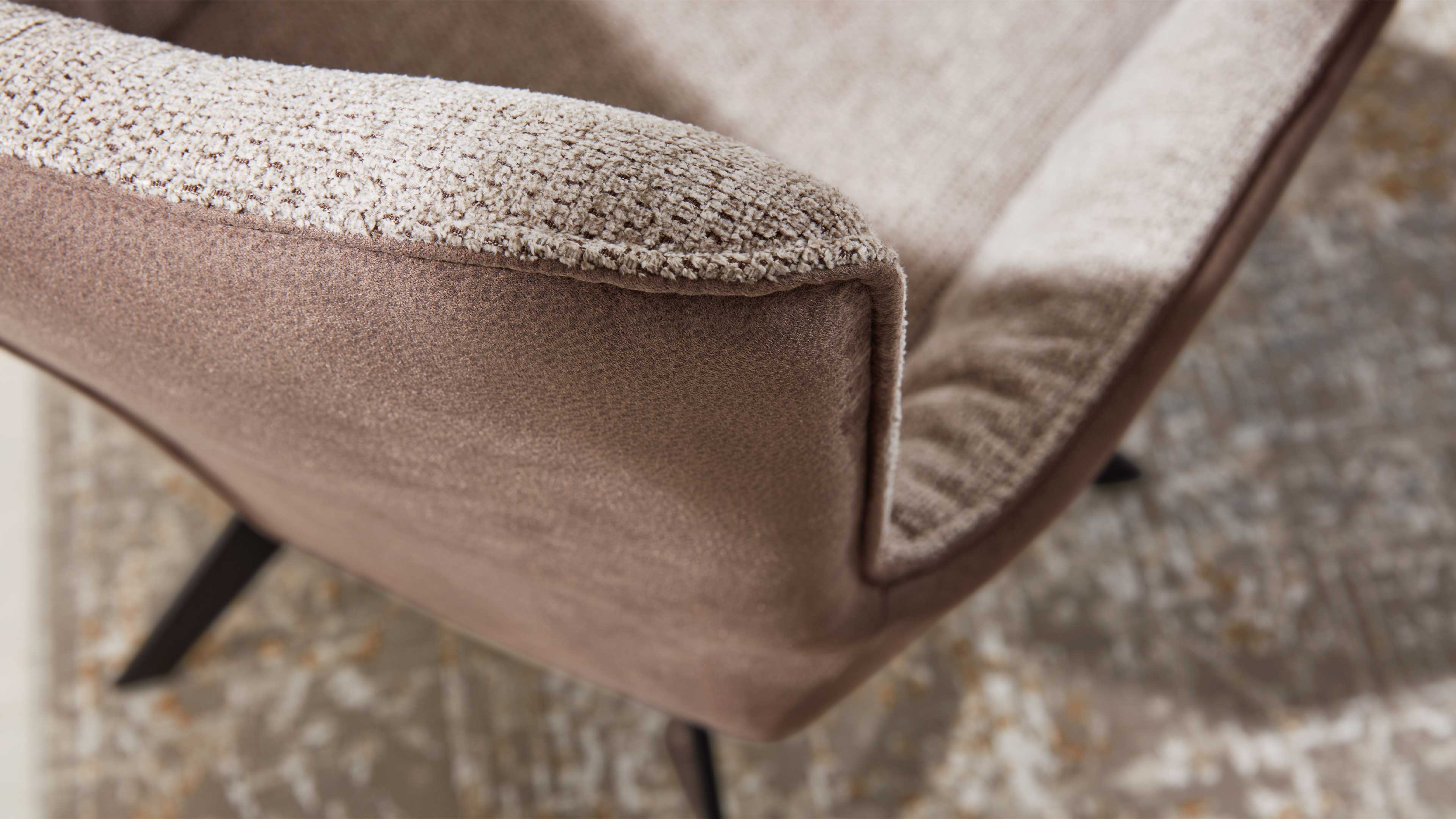 Nahaufnahme einer Sesselarmlehne mit strukturiert-beiger Polsterauflage, braunem Veloursaußenbezug, schwarzem Metallfuß und Musterteppich.