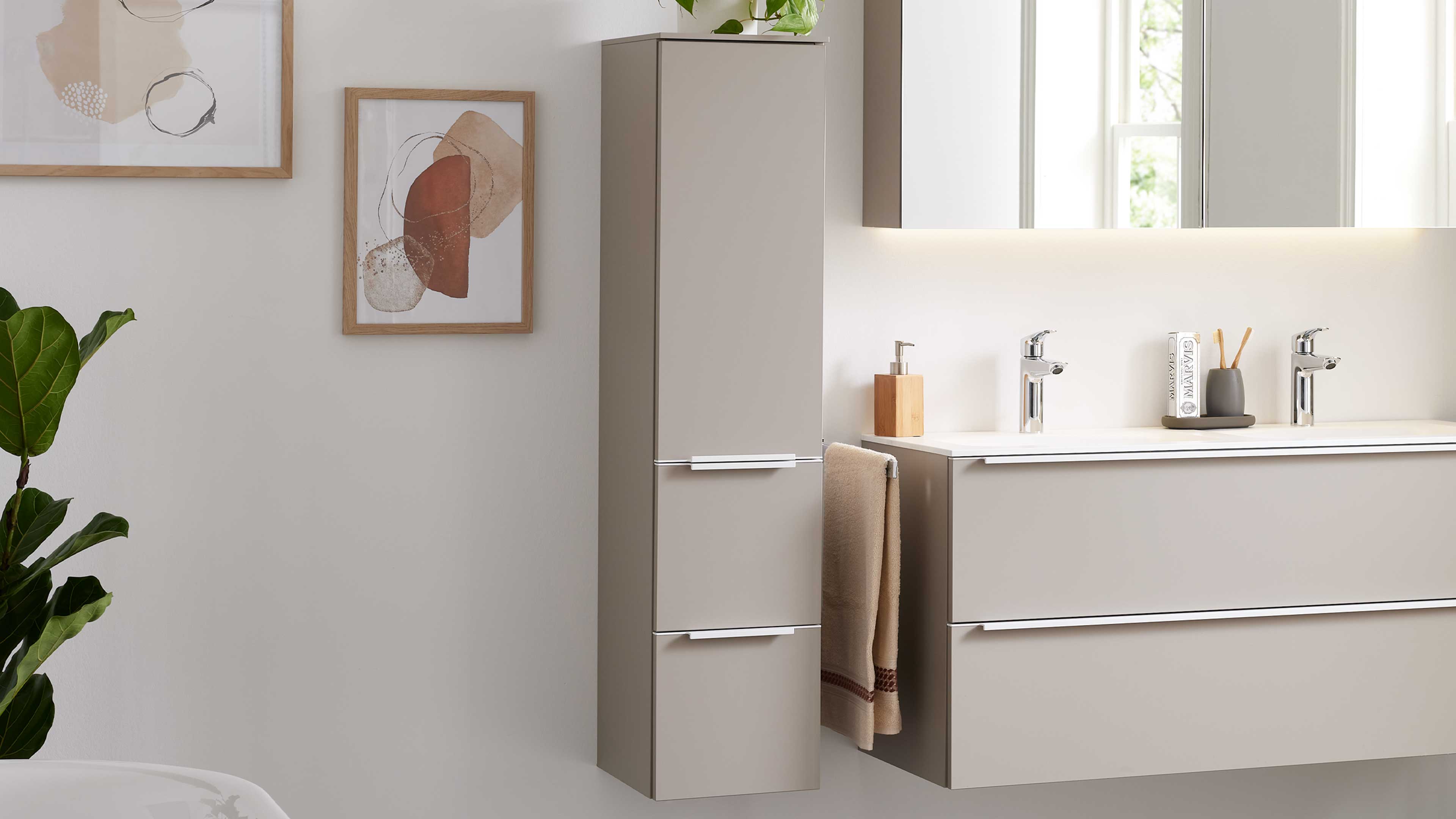 Hochhängender beiger Badschrank mit zwei Metallgriffen neben Waschtisch mit zwei Chromarmaturen, Seifenspender, Handtuch und Pflanze.