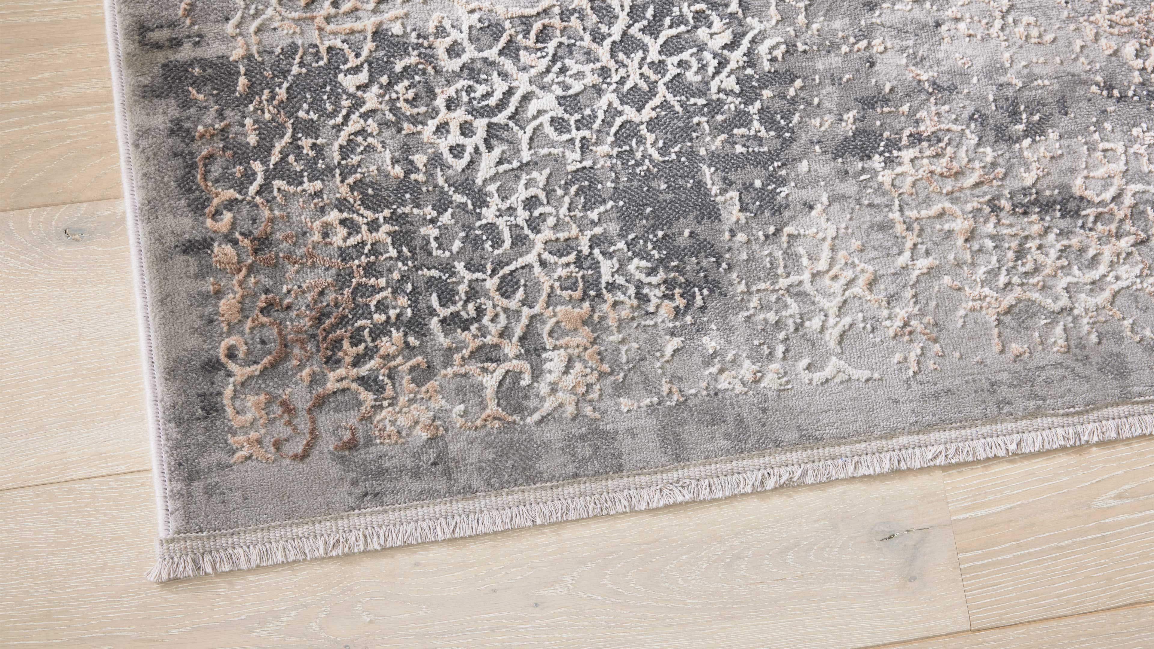 Eckansicht eines grauen Teppichs mit beigefarbenen reliefartigen Ornamenten und kurzer Fransenborte auf hellem Holzfußboden.