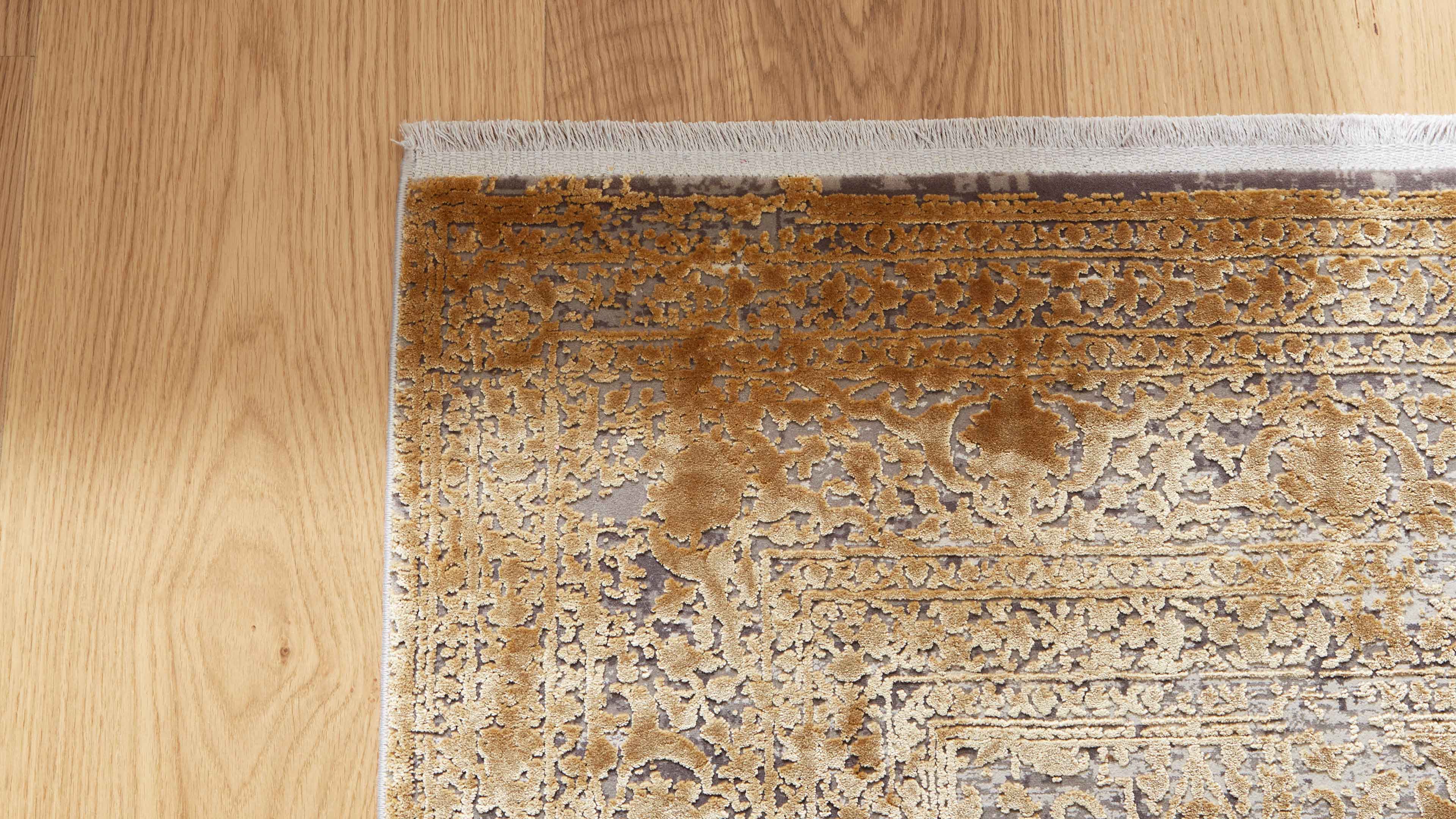 Eckansicht eines Teppichs mit goldbraunem ornamentalem Muster und weißen Fransen auf hellem Holzfußboden.