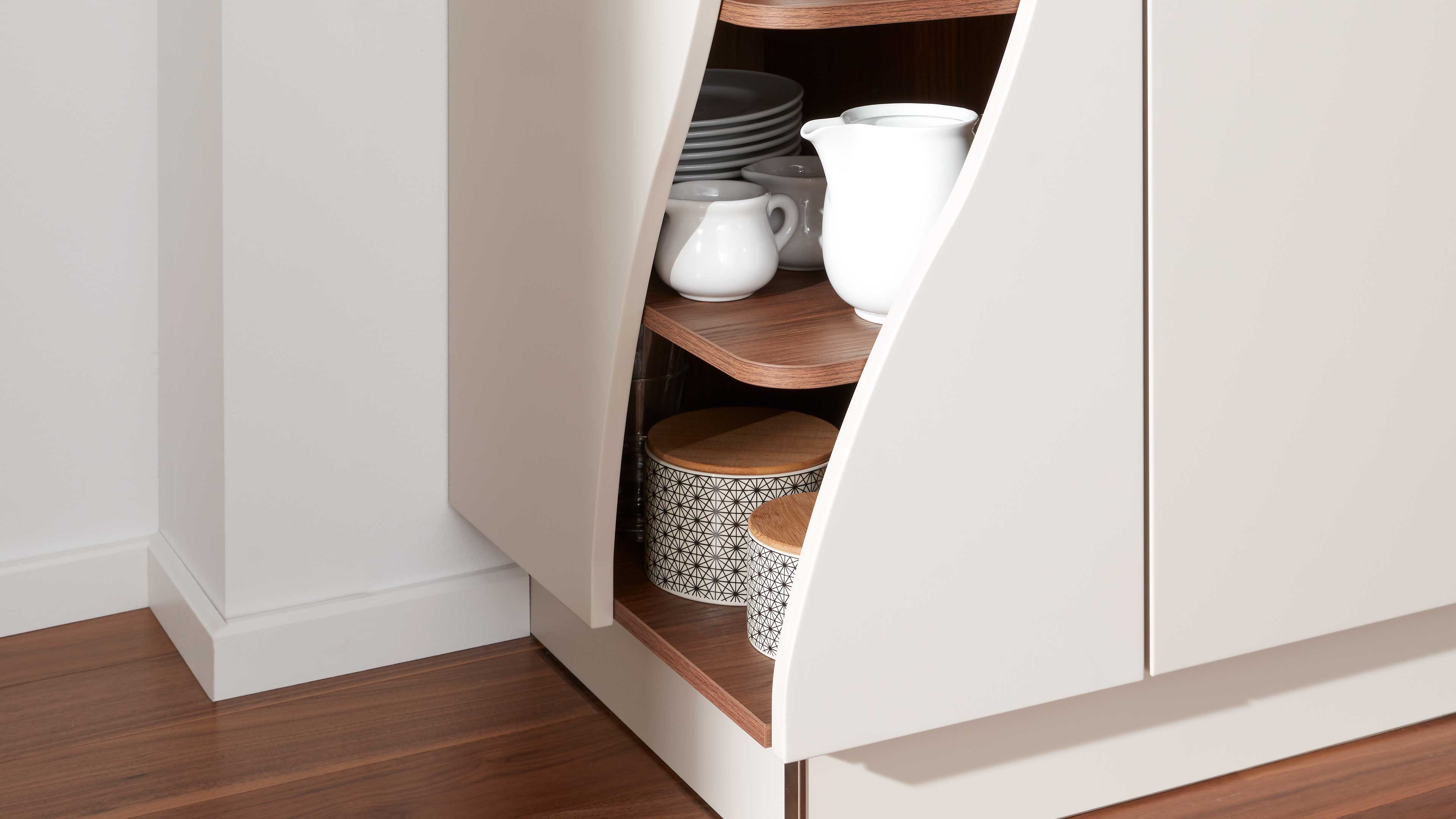 Teil eines matten cremefarbenen Küchenschranks mit geschwungener Öffnung, Holzregalen, weißer Keramikkanne, Tellern und gemusterten Dosen.