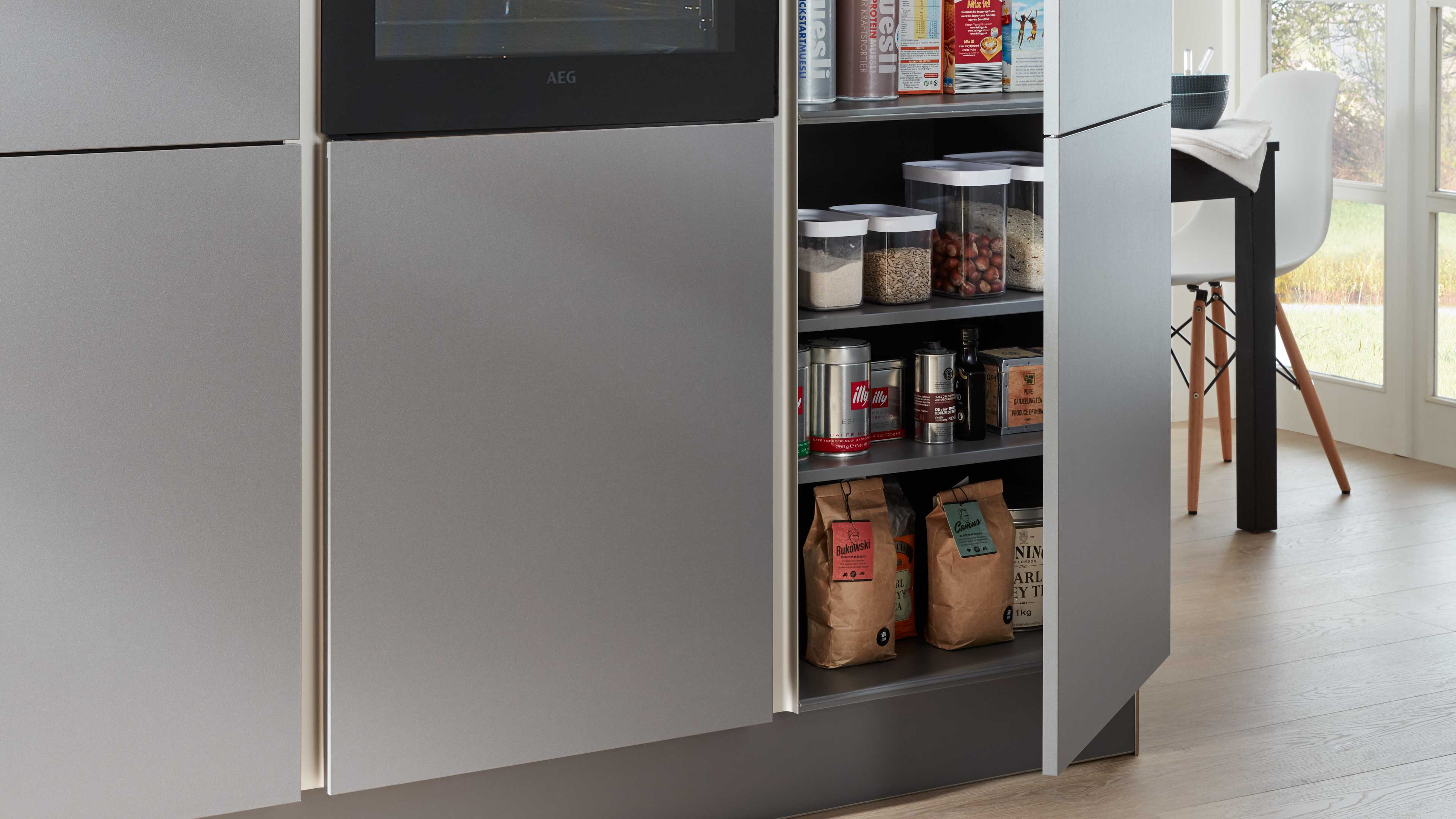 Offener schmaler Auszugsschrank mit grauen matten Fronten und eingelassener Griffkante, bestückt mit Vorratsdosen, Dosen und Papierbeuteln.