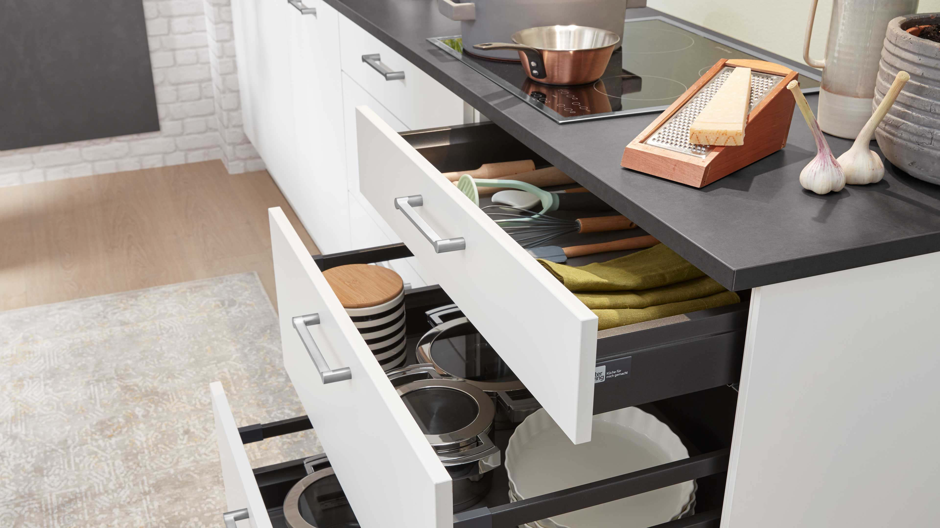Weiße Küchenfront mit drei offenen Schubladen, silbernen Griffen und dunkler Arbeitsplatte, darin Töpfe, Teller und Küchenutensilien.