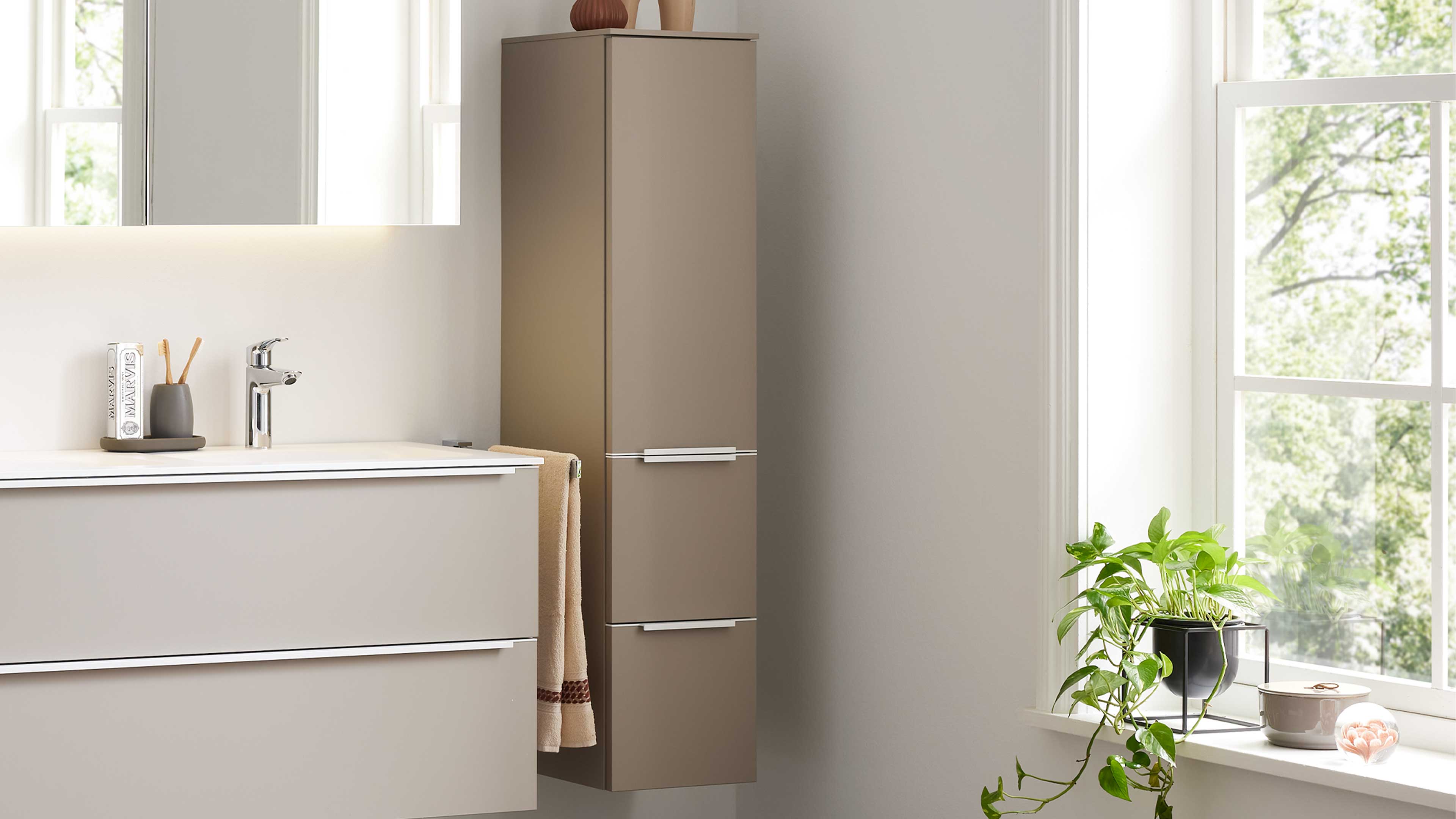 Schlanker beiger Hochschrank mit zwei silbernen Griffen neben Waschtischunterschrank mit Chromarmatur und hängendem Handtuch.