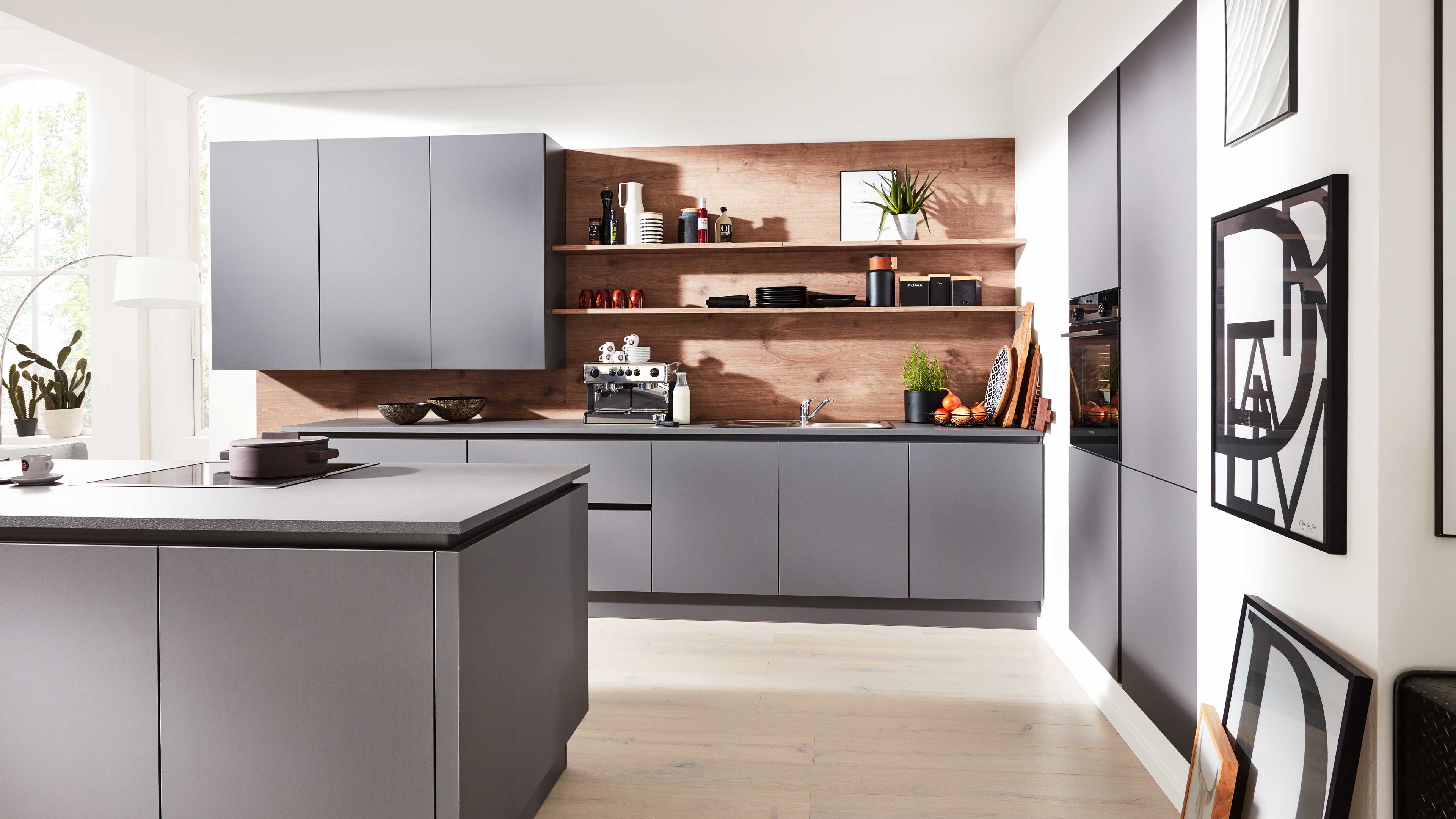 Graue Kücheninsel und Wandschränke vor Holzrückwand mit offenen Regalen, Espressomaschine, Spüle, Pflanzen und Wandbildern.