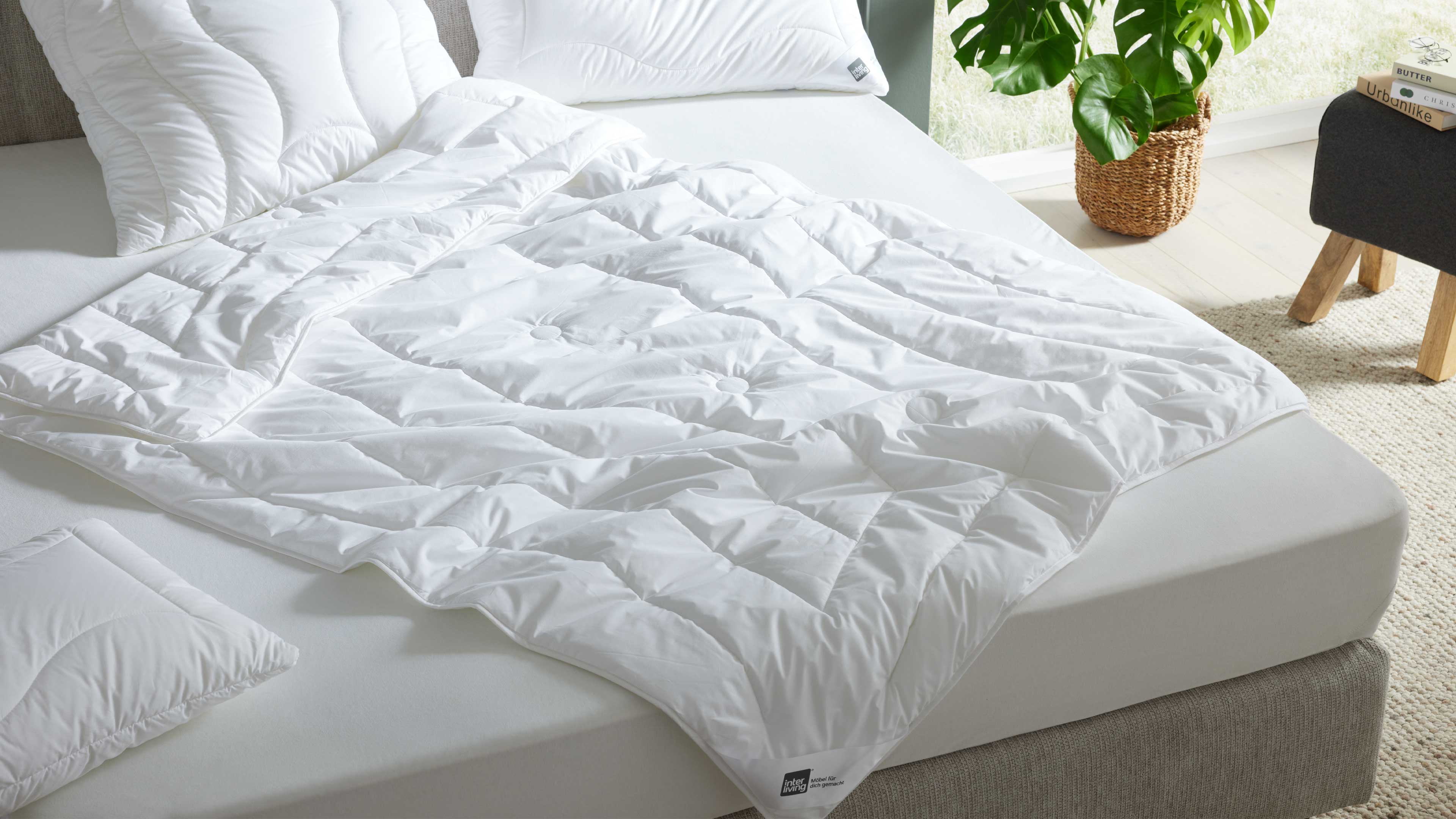 Weiße gesteppte Bettdecke liegt auf Matratze mit zwei weißen Kissen, grauem Bettrahmen, Korb mit Grünpflanze und Hocker.