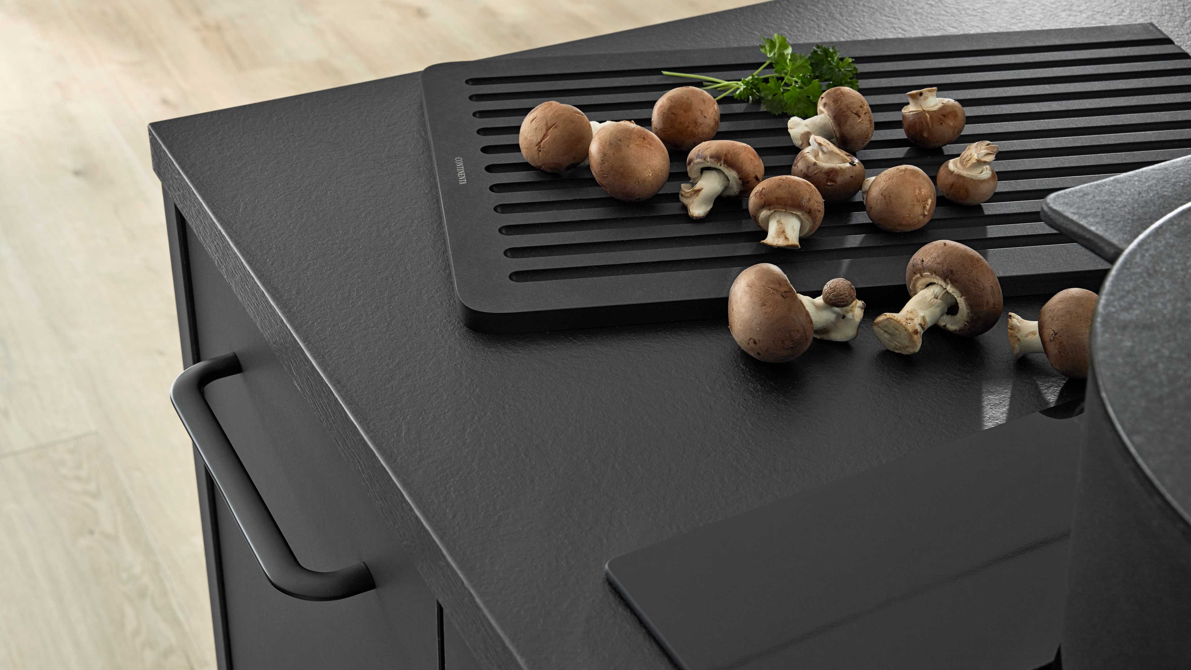 Schwarze Küchenarbeitsplatte mit schwarzem Griff, darauf ein geschlitztes Schneidebrett, braune Champignons und etwas Petersilie.