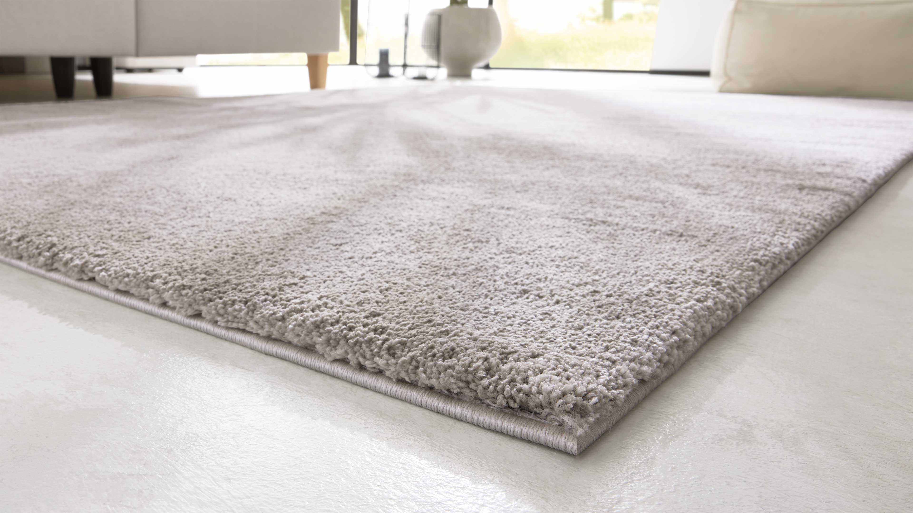 Hellgrauer, dichter Teppich mit abgerundeter Kante liegt auf hellem Boden; im Hintergrund Sofabeine und Vase.