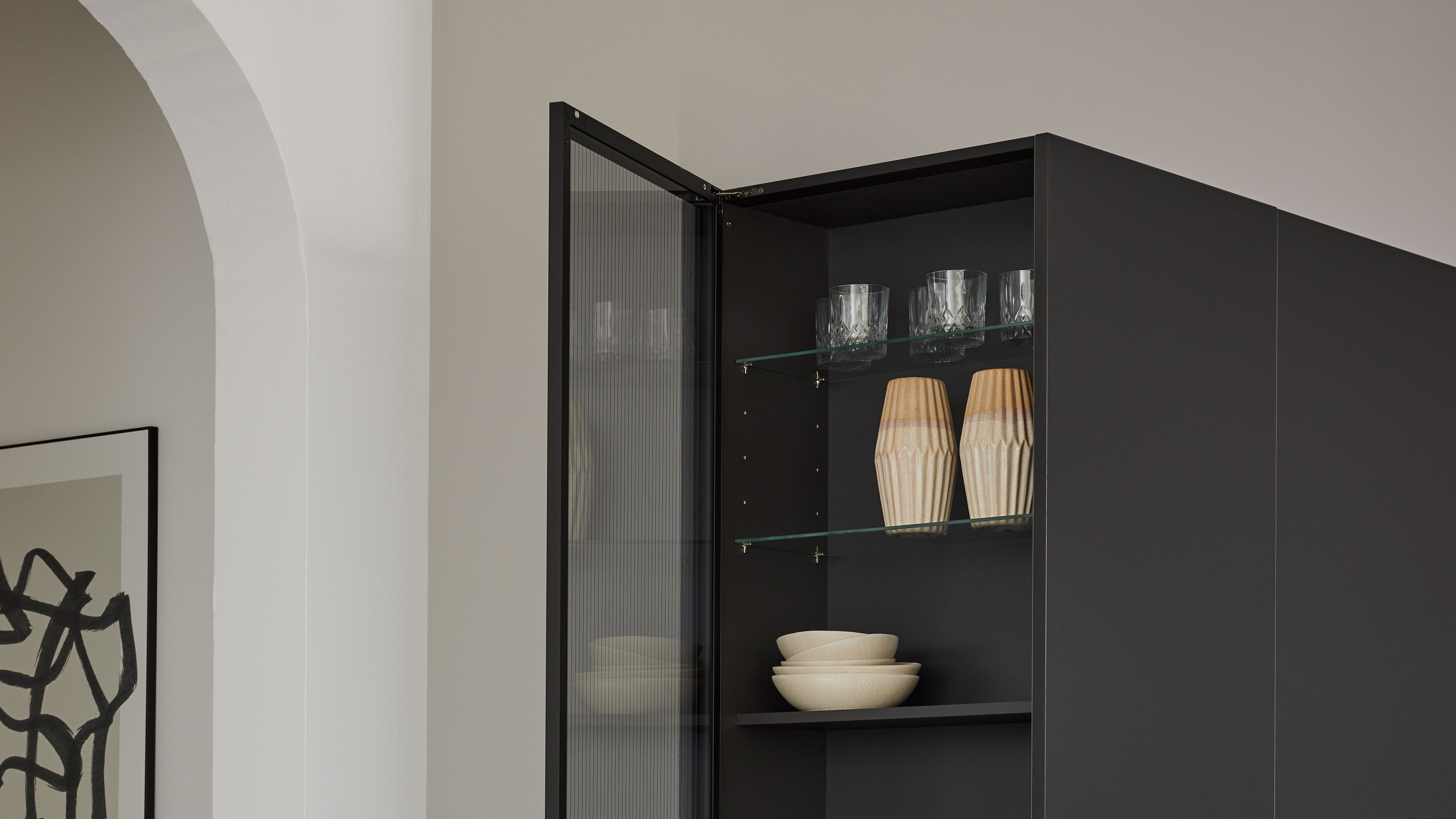 Schwarzer Hochschrank mit geöffneter Glastür; innen Glasböden mit mehreren Trinkgläsern, zwei Holzvasen und gestapelten beigen Schalen.