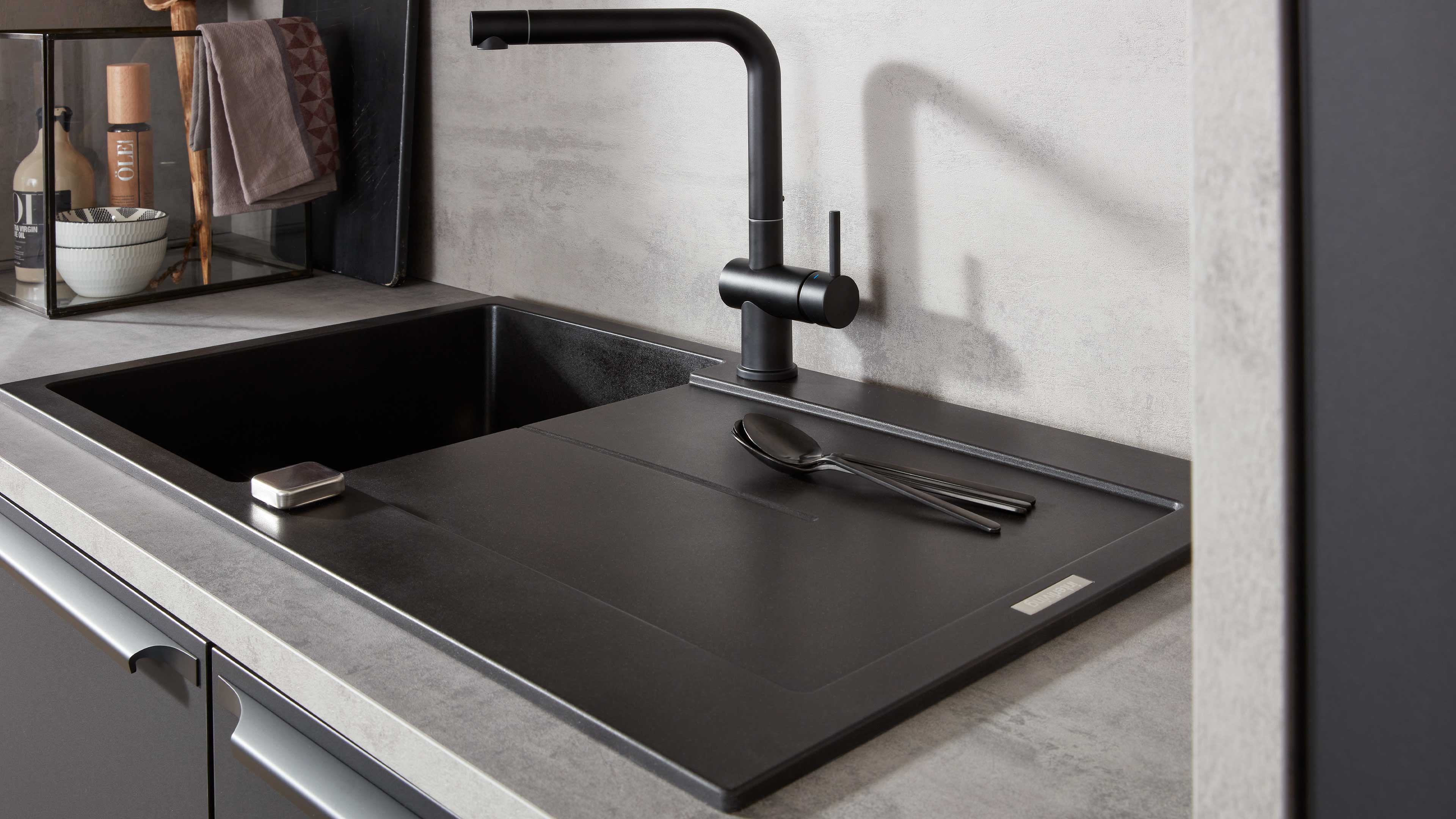 Schwarze Küchenspüle mit aufgelegter Abtropffläche, schwarzem Einhebelmischer, zwei Löffeln und einer Glasvitrine mit Schalen links.