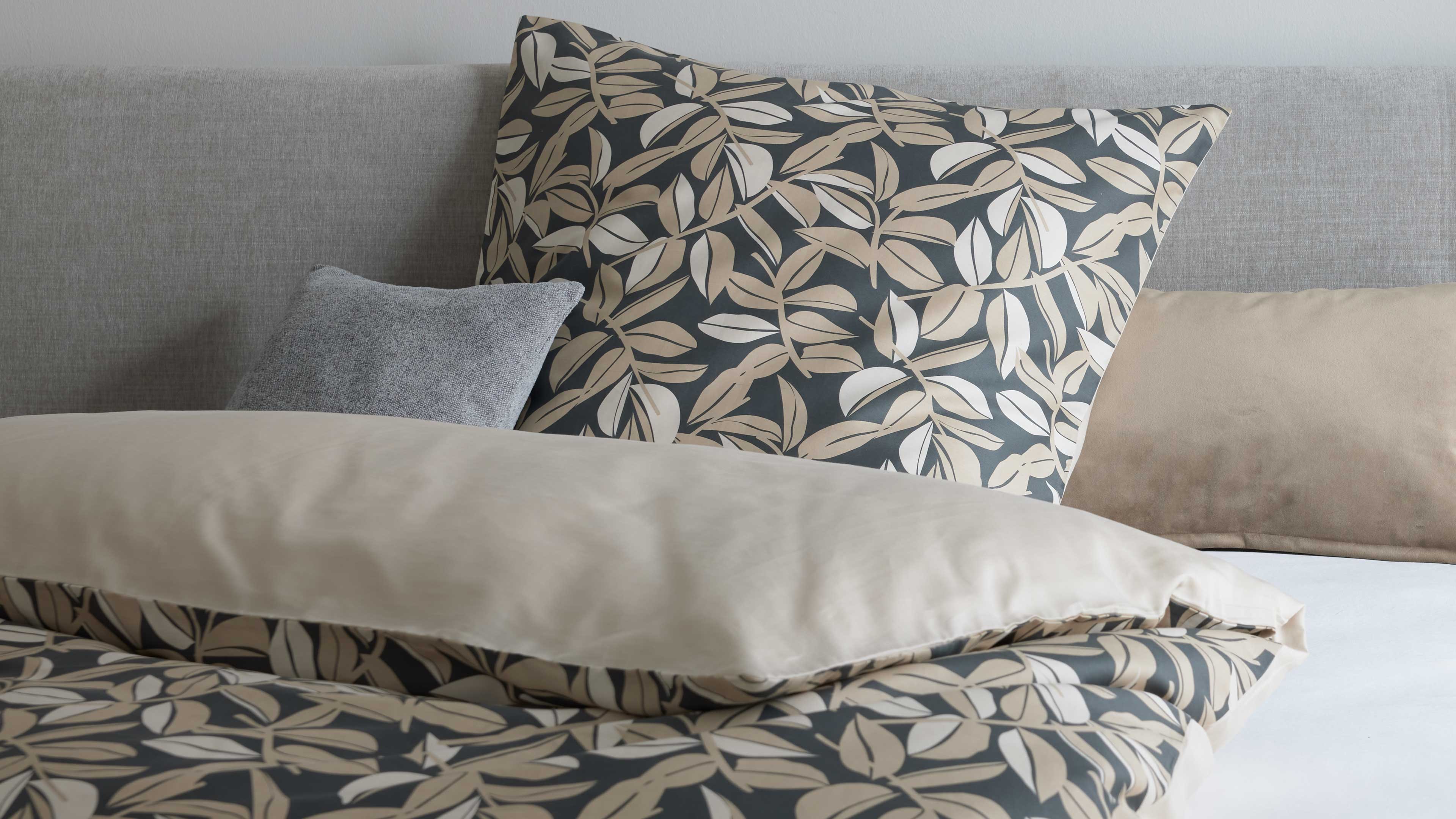 Großes Kissen mit dunkelgrauem Hintergrund und beige-weißen Blattmotiven vorn, beigefarbene Bettdecke und graues Zierkissen am Kopfteil.