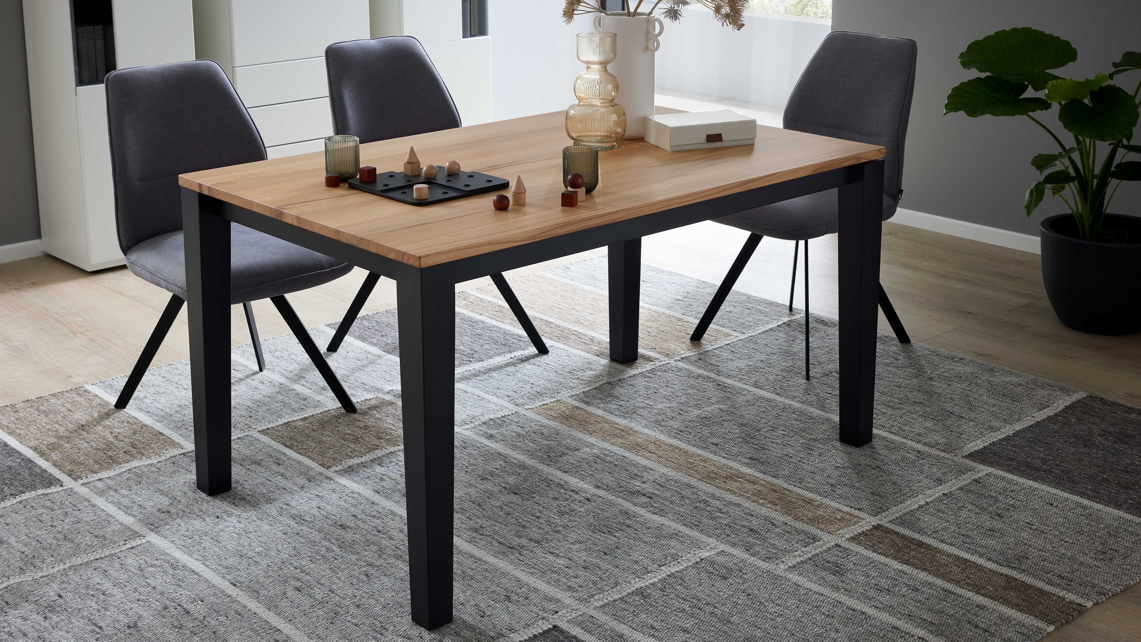 Esstisch mit hellbrauner Holzplatte und schwarzen Metallbeinen, drei graue Polsterstühle, Vasen, Spielsteine und Teppich.