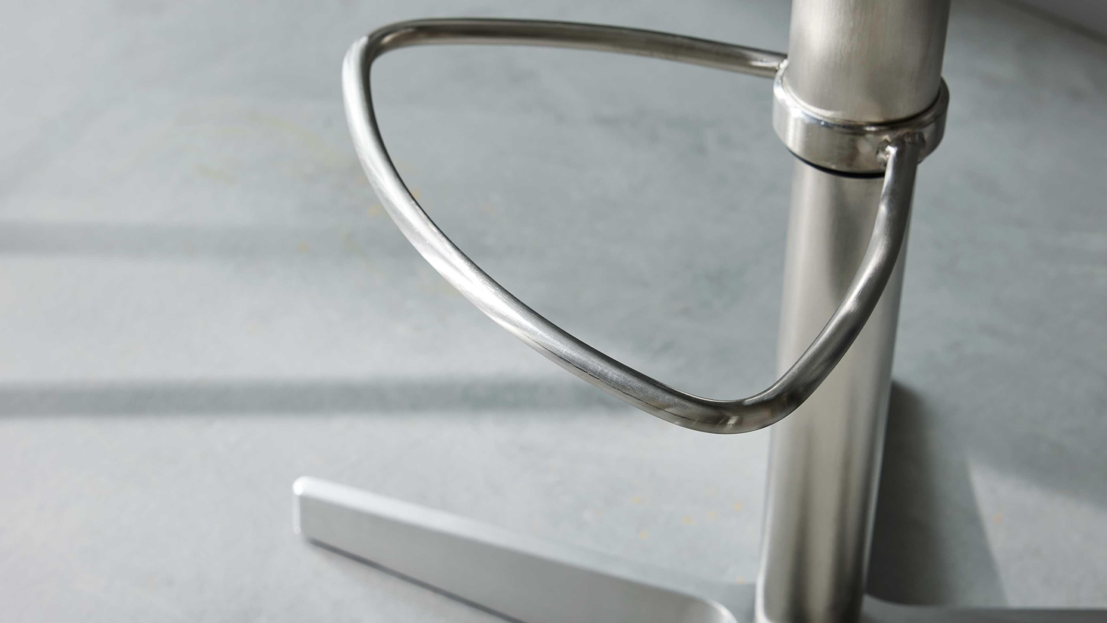 Gebogener, dreieckiger Fußring aus gebürstetem Metall an einer runden Stuhlsäule mit kreuzförmigem Metallfuß auf grauem Boden.