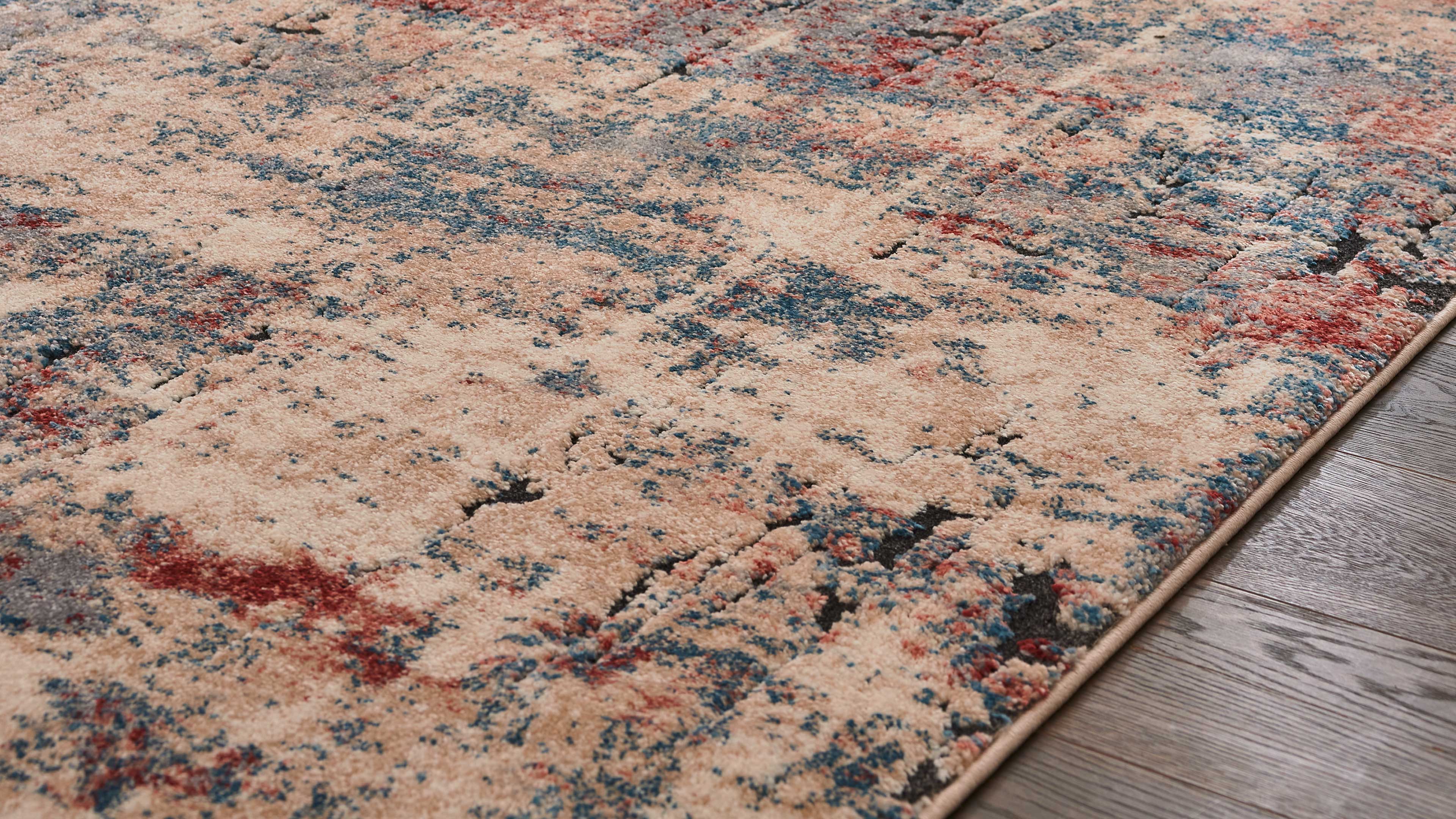 Beiger Teppich mit flächigem Muster in Blau, Rot und Schwarz, sichtbarer Florstruktur und schmaler Einfassung auf Holzboden.