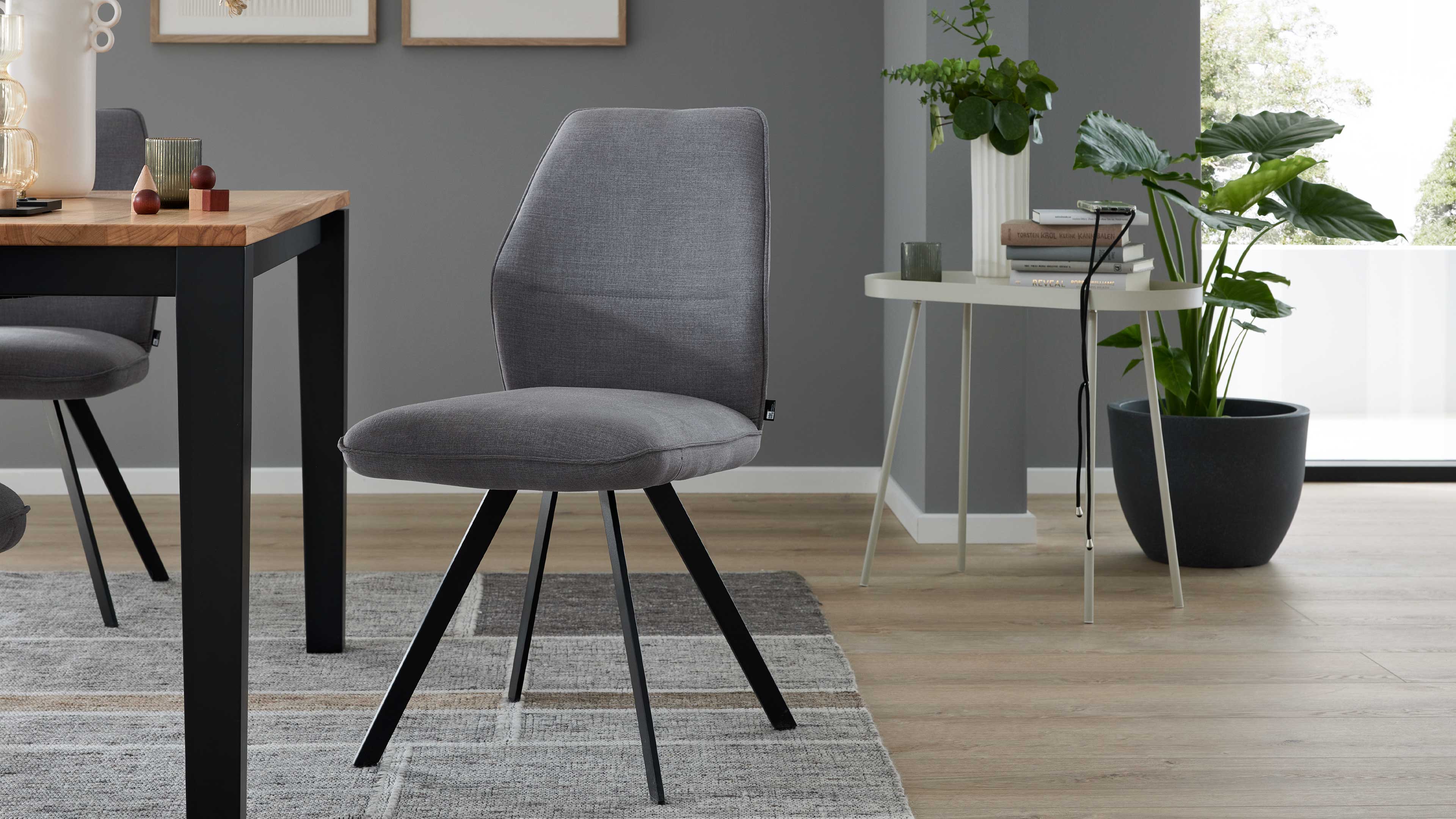 Grau bezogener Stuhl mit gepolstertem Sitz und schwarzen Metallbeinen auf Teppich, daneben Holztisch und Beistelltisch mit Pflanze.
