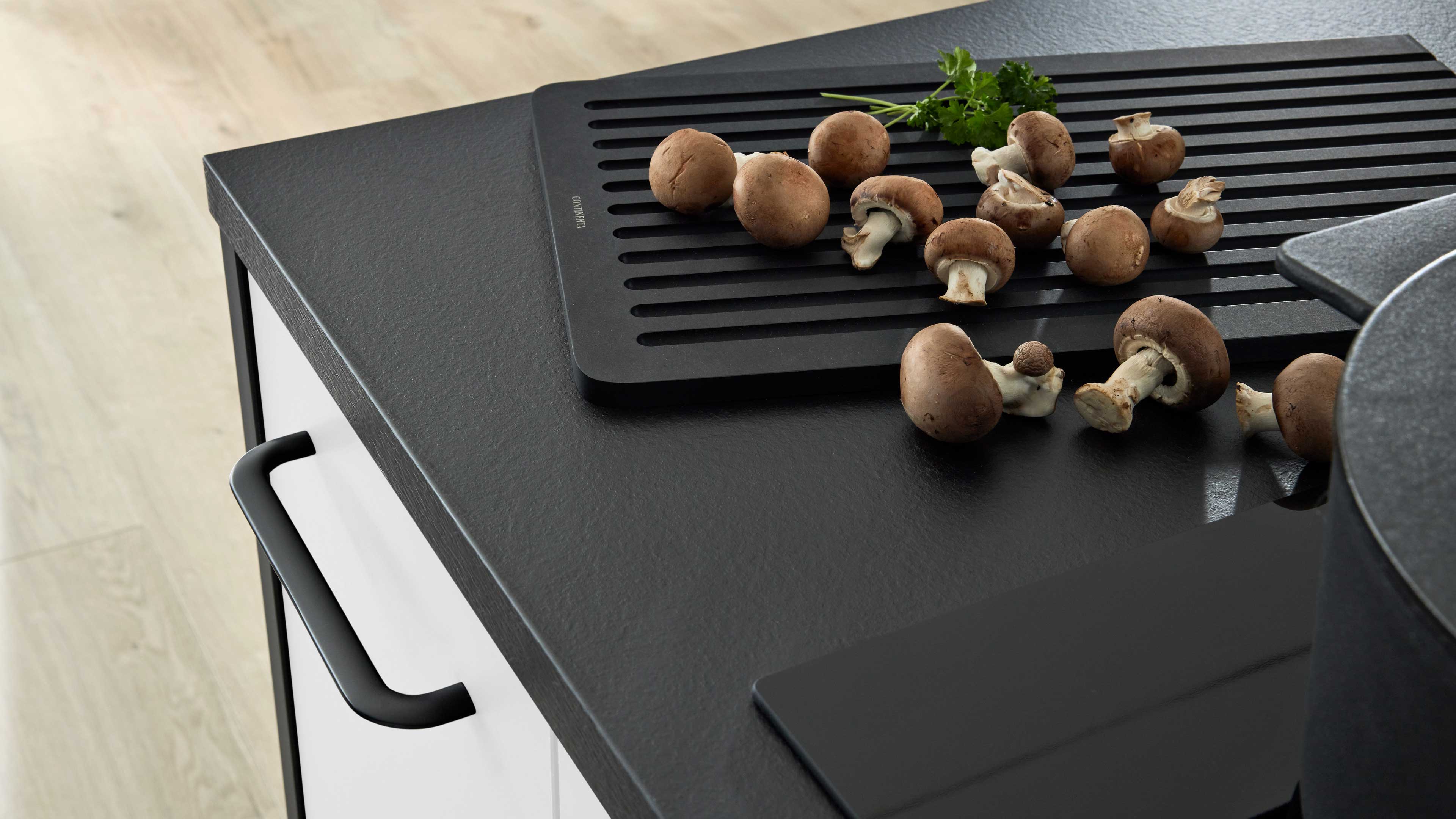 Schwarze Arbeitsplatte mit schwarzem, längs geriffeltem Abtropfbrett und braunen Champignons, links weiße Schublade mit schwarzem Griff.
