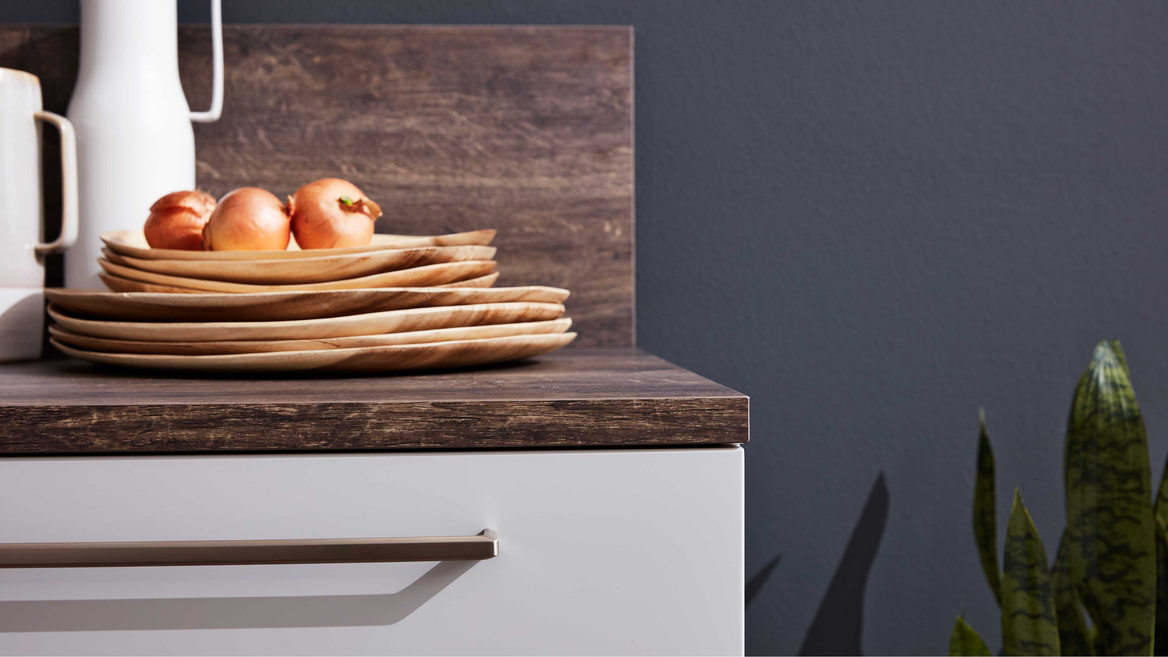 Gestapelte Holzplatten mit Zwiebeln auf dunkler Holzarbeitsplatte, links eine weiße Kanne, darunter eine weiße Schublade mit Metallgriff.