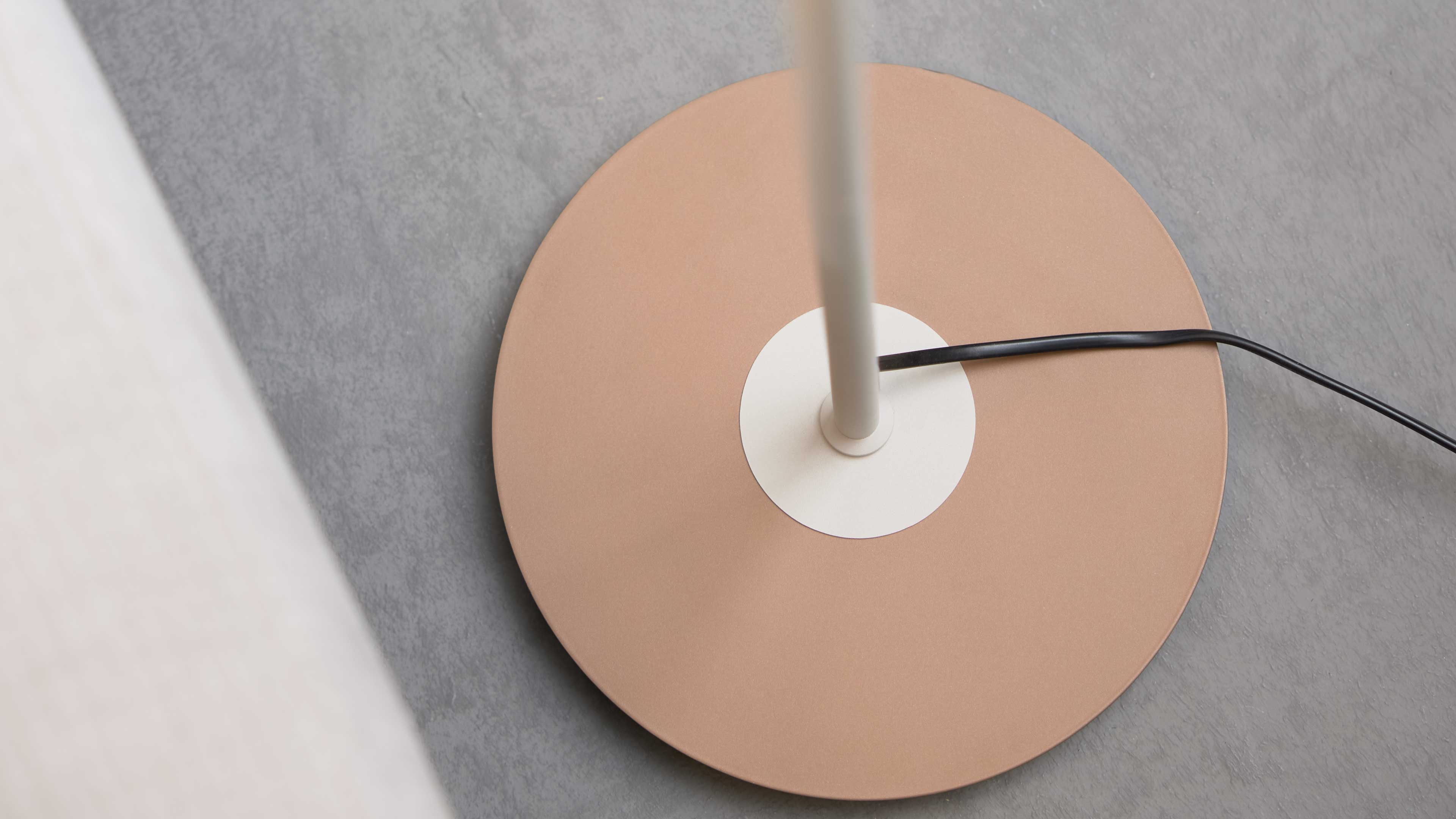 Runder roségoldener Lampenfuß mit weißer Mittelplatte und aufgesetztem Stab, schwarzes Kabel quer auf grauem Boden.
