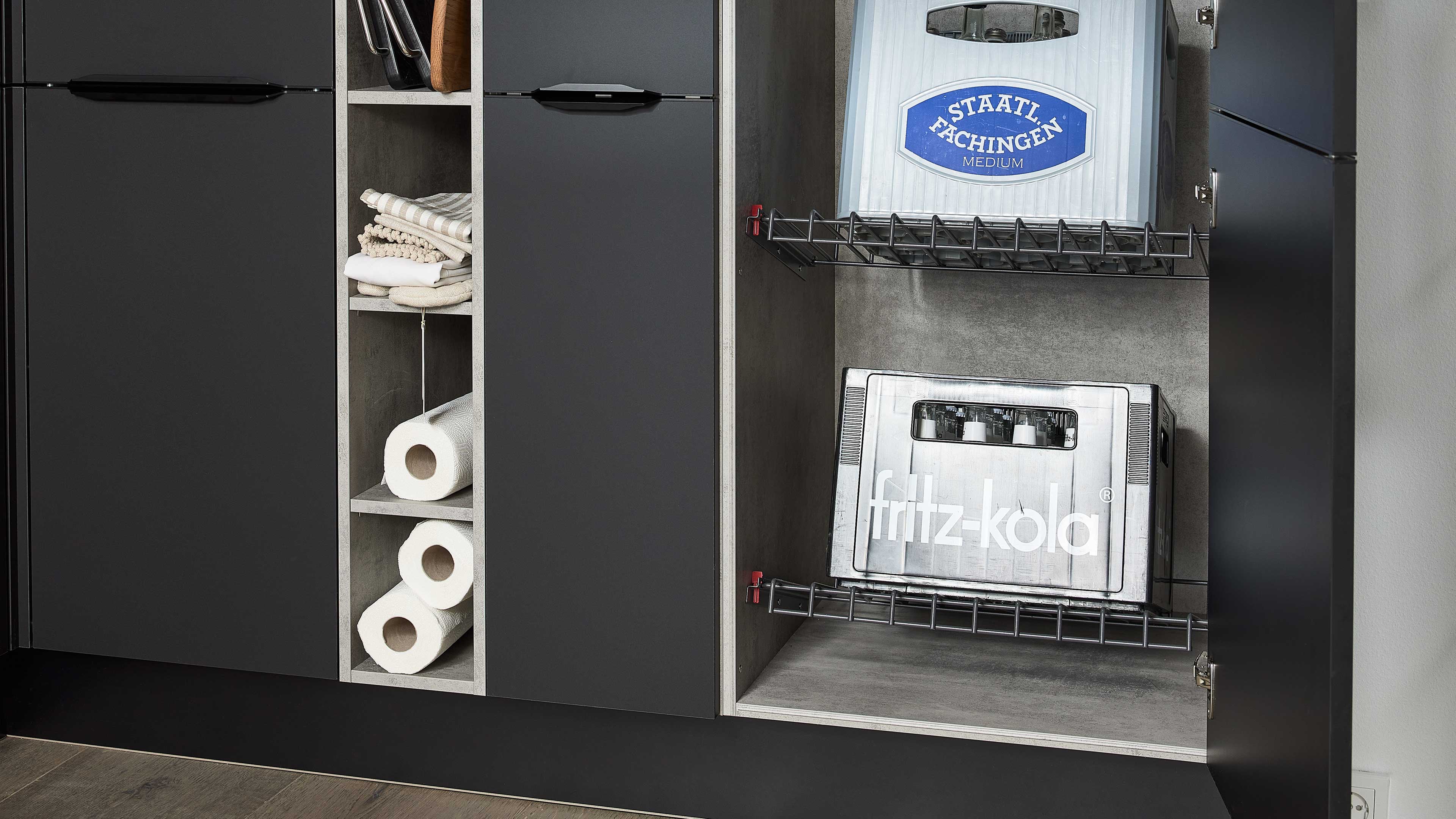 Offener Unterschrank mit Drahtgitterschubladen, unten Fritz-Kola-Kiste, oben Staatl. Fachingen; links Fächer mit Tüchern und Rollen.