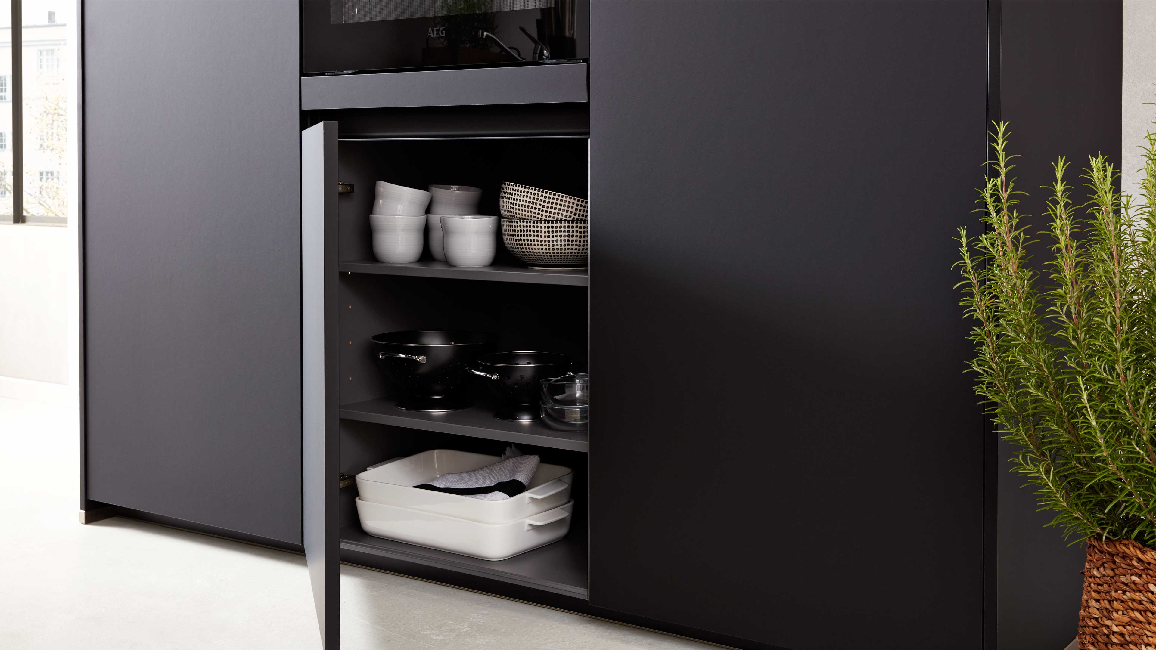 Offenes dunkelgraues Küchenmöbel mit Regalfächern, darin weiße Schalen, gemusterte Schüssel, schwarze Töpfe, Auflaufformen und Topfkräuter.