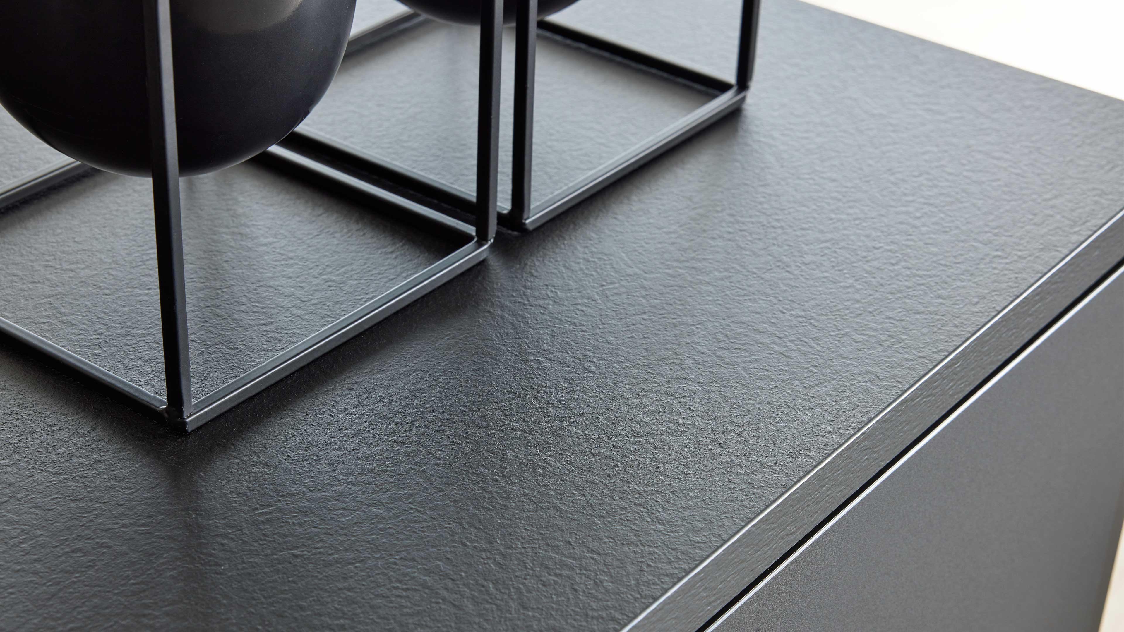 Schwarze, leicht strukturierte Oberseite eines Schranks mit zwei quadratischen Metallgestellen und Teil eines runden Topfes.