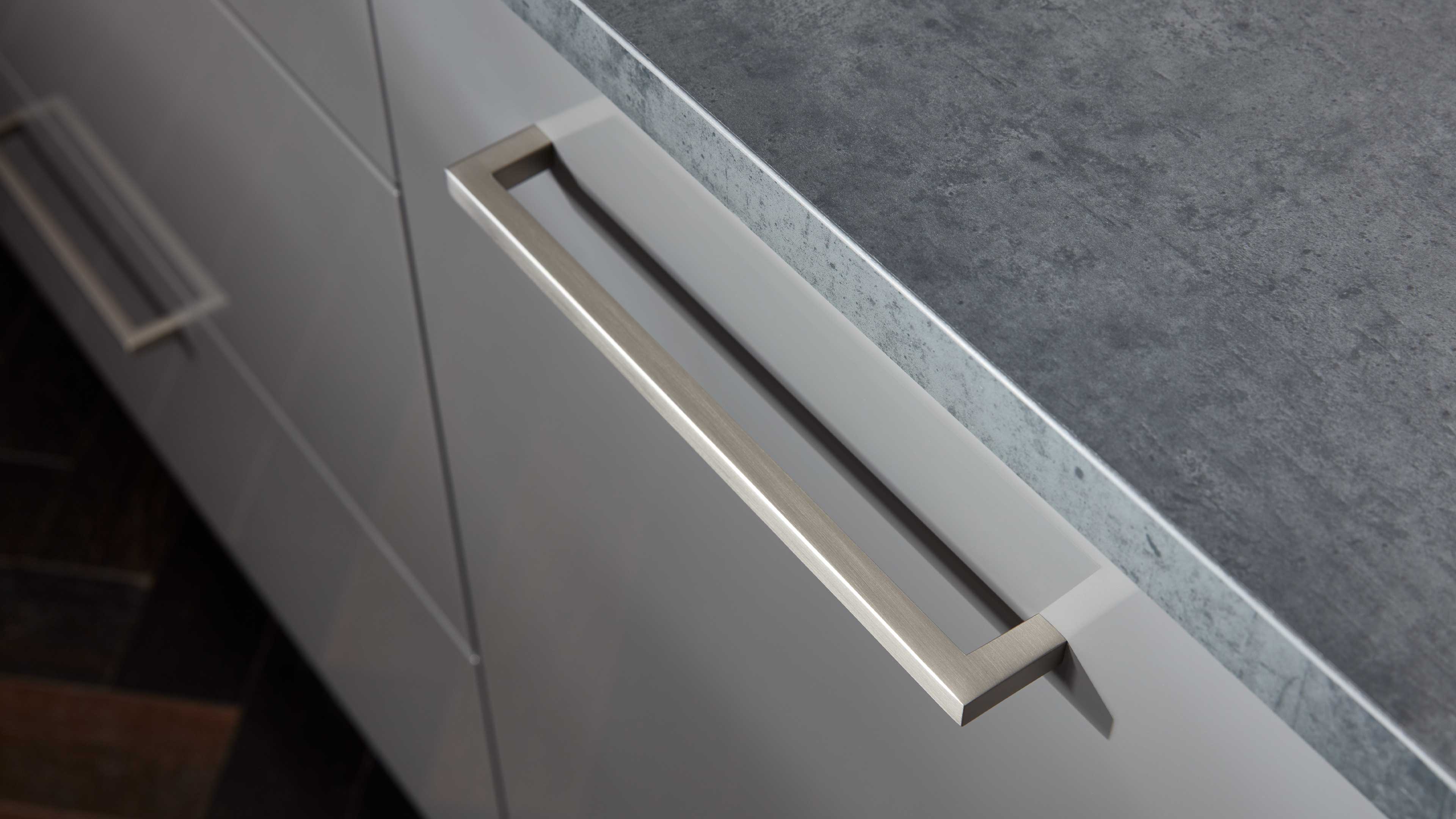 Küchenschrank-Schubkastengriff in rechteckiger Form aus gebürstetem Metall an heller Front unter grauer Steinoptik-Arbeitsplatte.