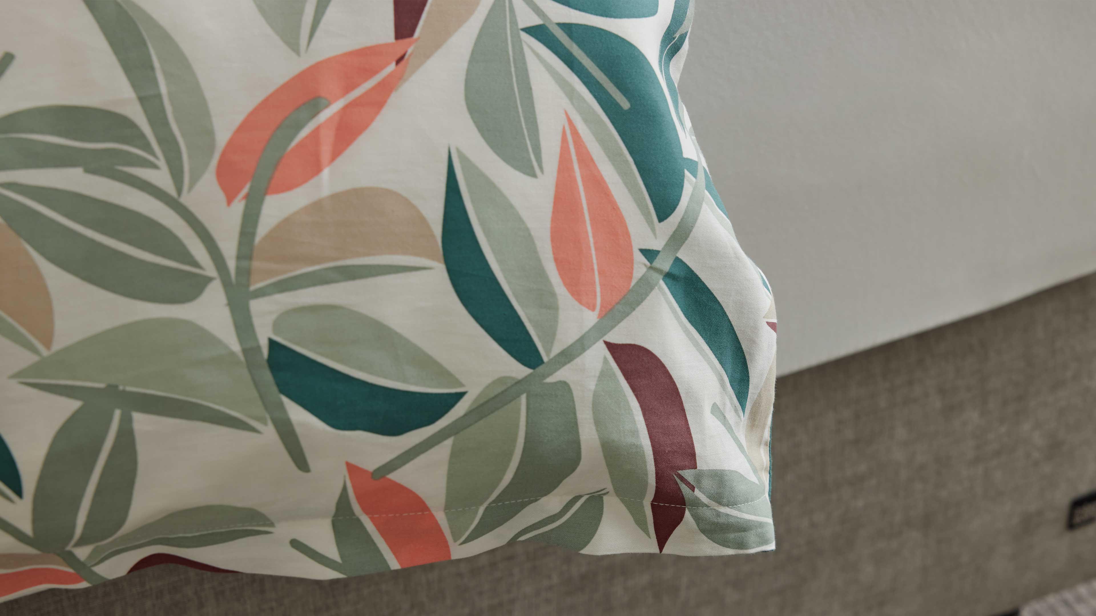 Ecke eines weißen Bettbezugs mit mehrfarbigem Blattmuster in Grün, Koralle und Beige, über einem grauen, gepolsterten Bettrand hängend.