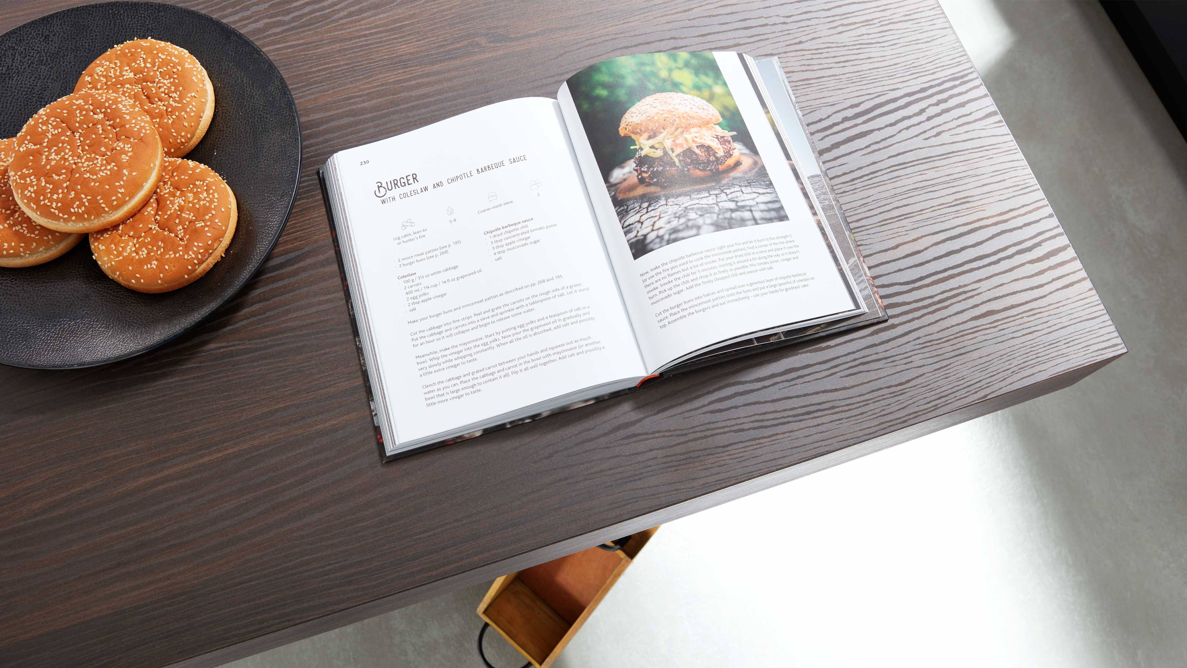 Dunkler Holztisch mit aufgeschlagenem Kochbuch (Burgerfoto), schwarzer Platte mit vier Sesambrötchen und geöffnetem Schubfach.