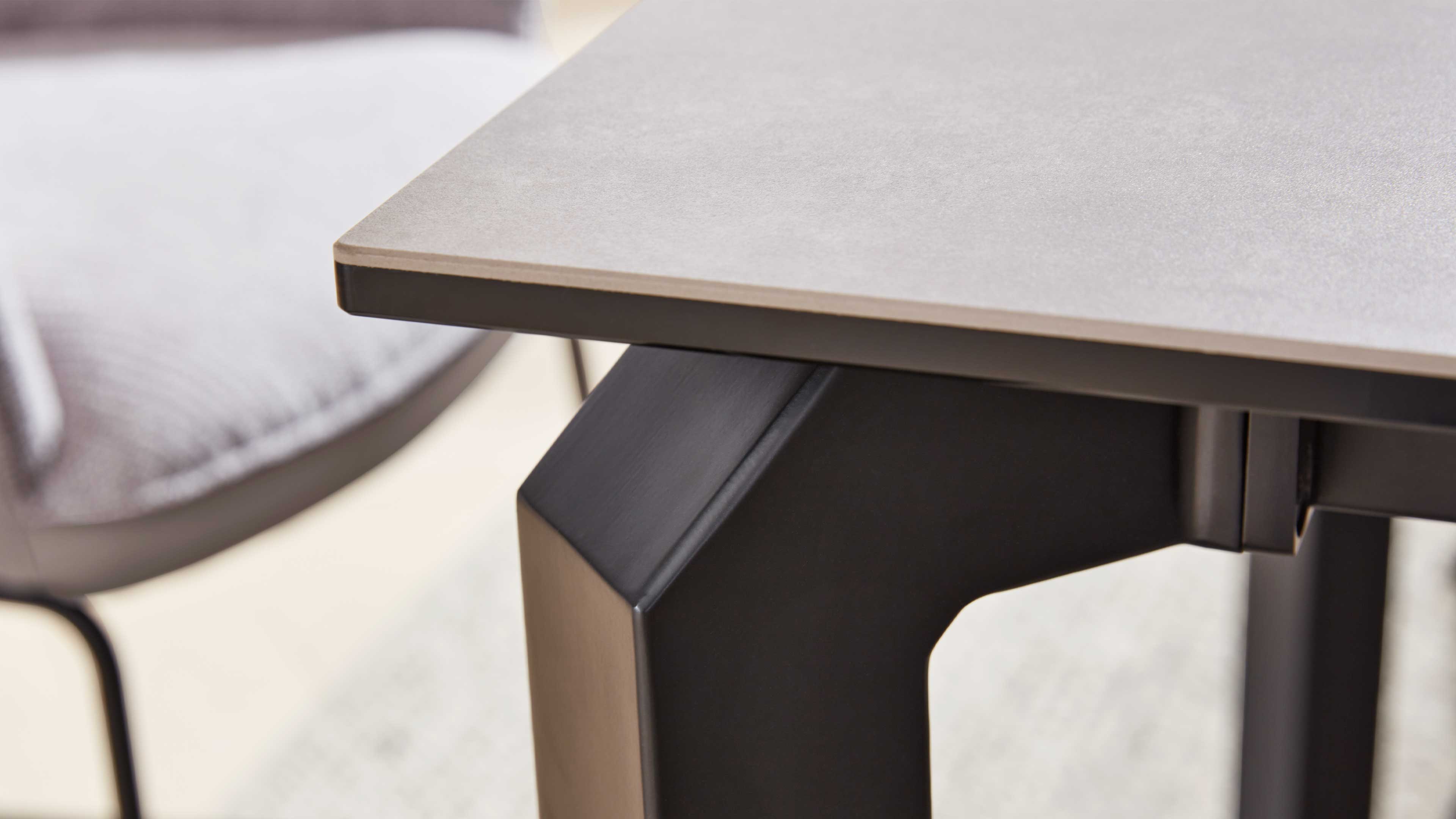 Tischplatte in hellgrauem Ton mit dünner Kante und schwarzem, kantigem Metallbein, links unscharf grauer Stuhl.