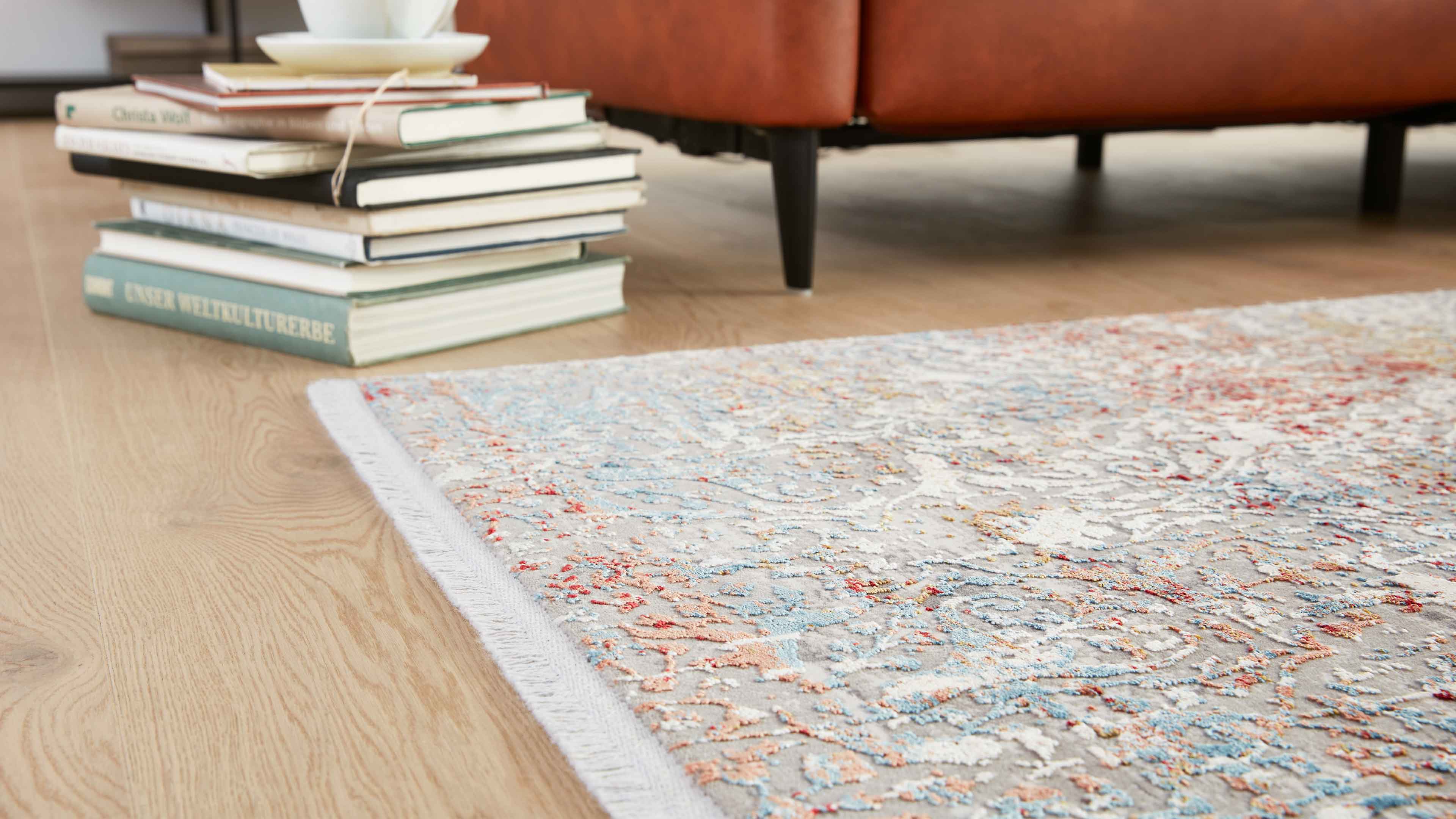 Teppich mit blau-orange-weißem Ornament und Fransen auf hellem Holzfußboden, im Hintergrund Bücherstapel und braunes Sofa mit schwarzem Bein.