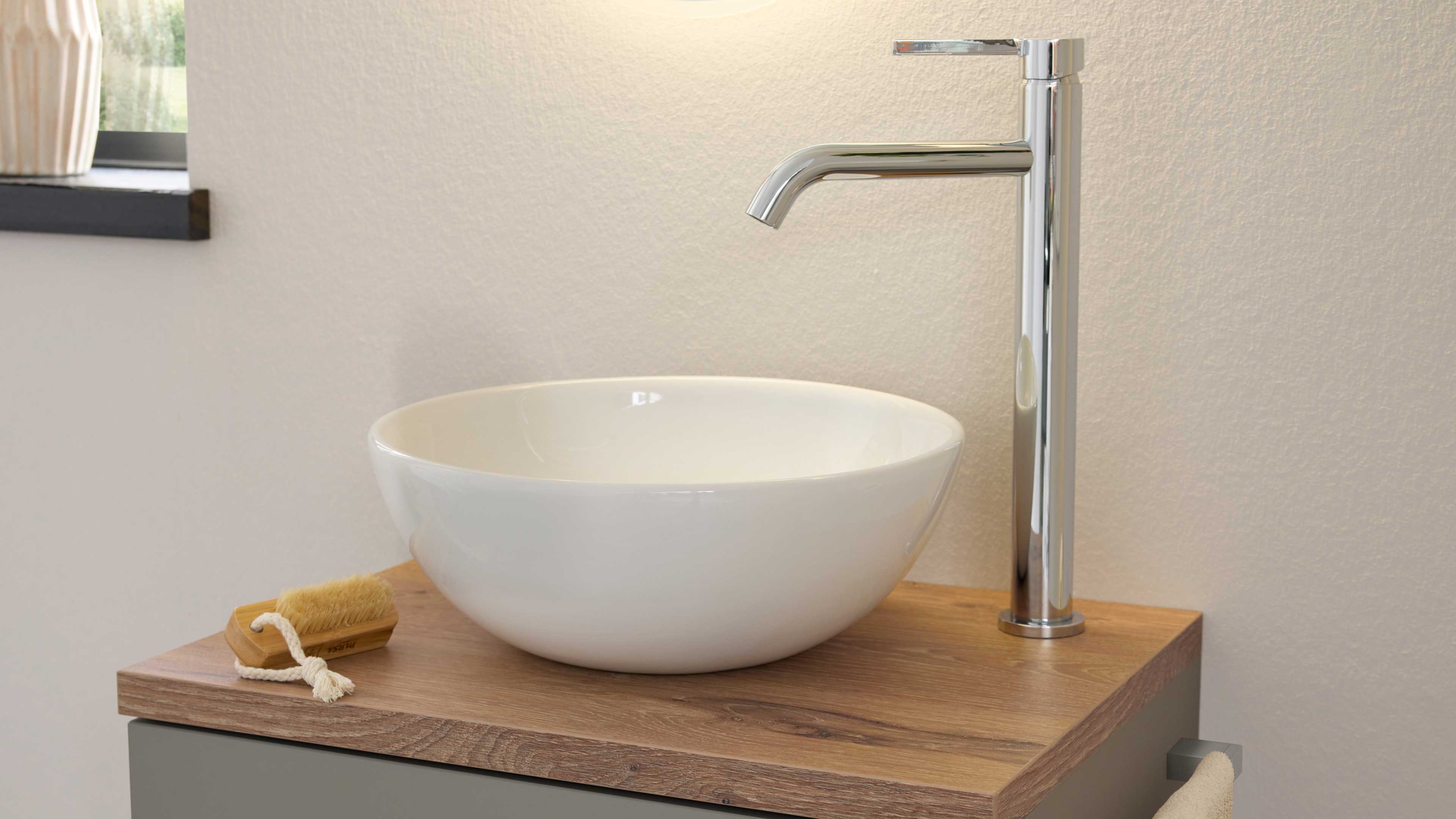 Runde weiße Aufsatzschale auf Holzplatte mit hohem verchromten Einhebelmischer, kleiner Bürste und Handtuch.