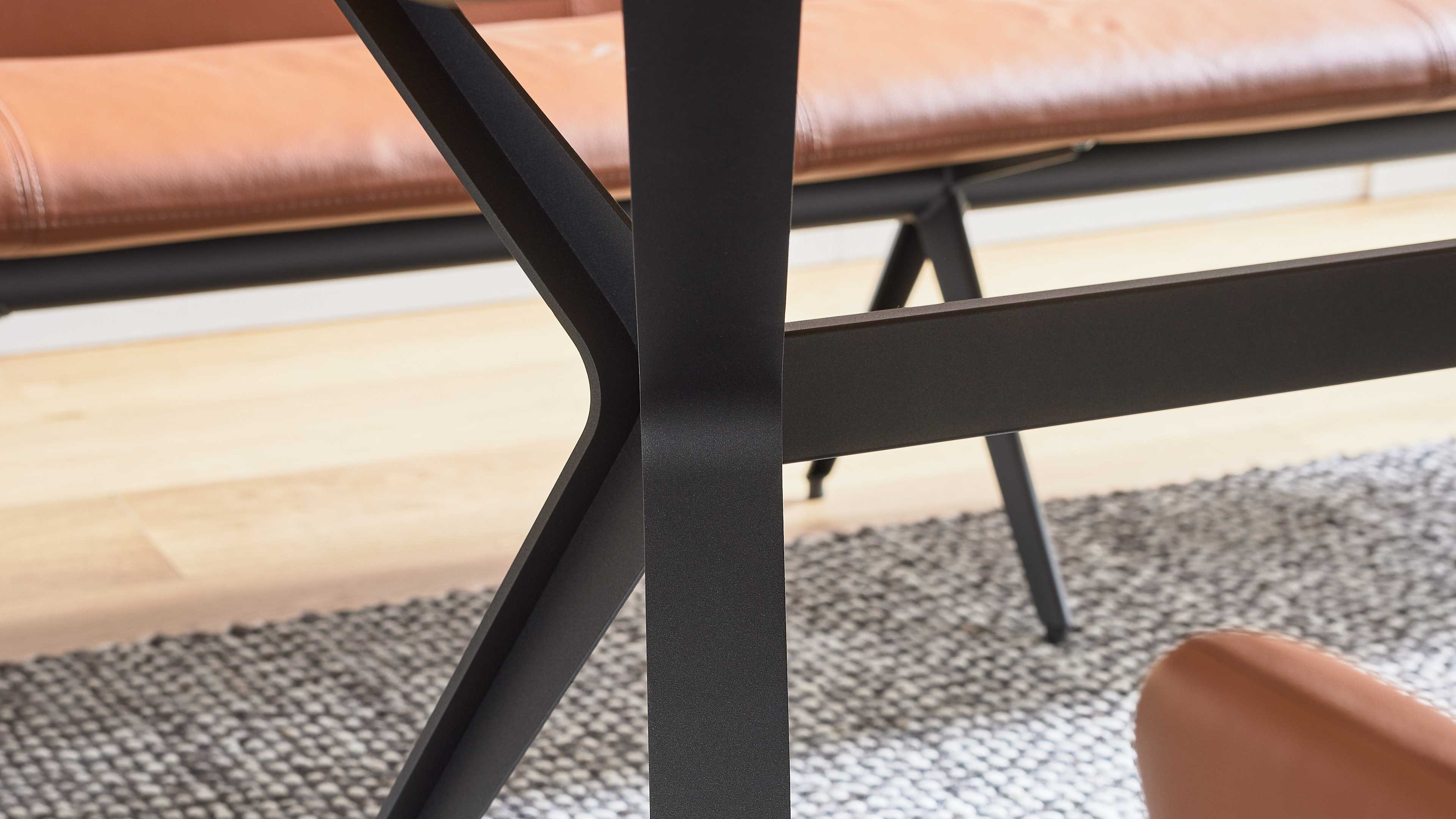 Schwarzes kreuzförmiges Metallgestell im Vordergrund, dahinter eine braune Lederbank und eine abgerundete Polsterkante.