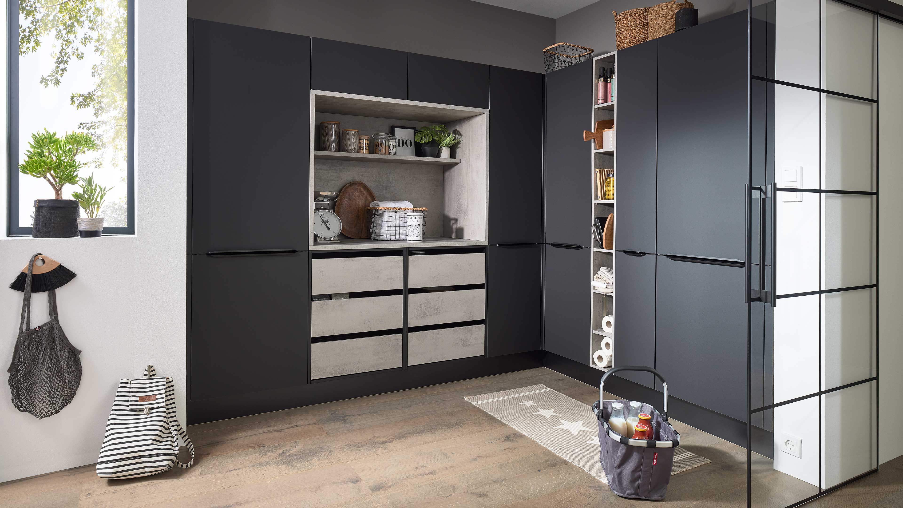 Dunkle Küchen- und Hochschränke in L-Form mit offenen Regalfächern und Sichtbeton-Fronten, Pflanzen am Fenster, Einkaufskorb.