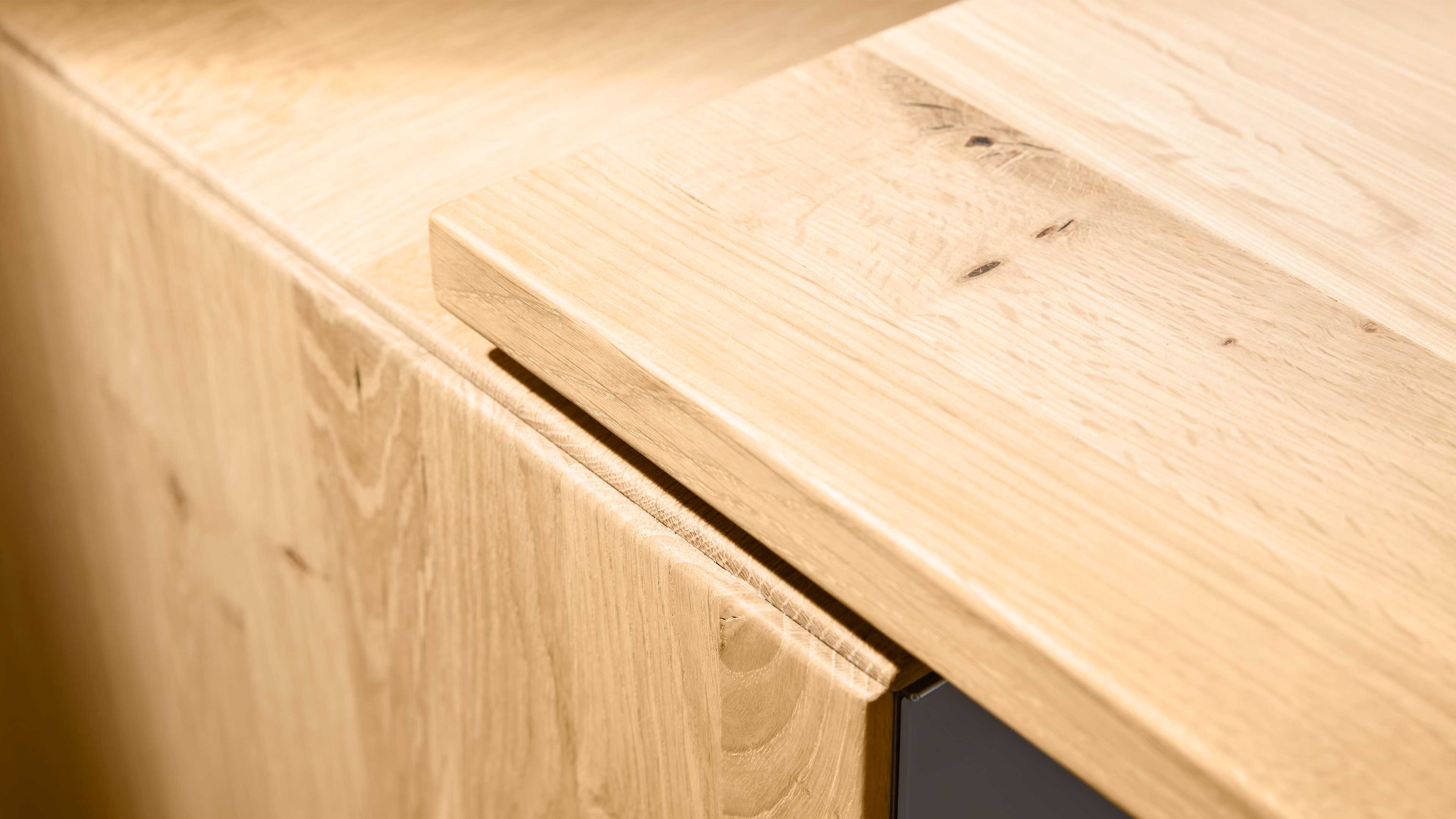 Abgeflachte Tischplatte aus hellem Eichenholz mit sichtbarer Maserung und Griffspalt über einer Schranktür.