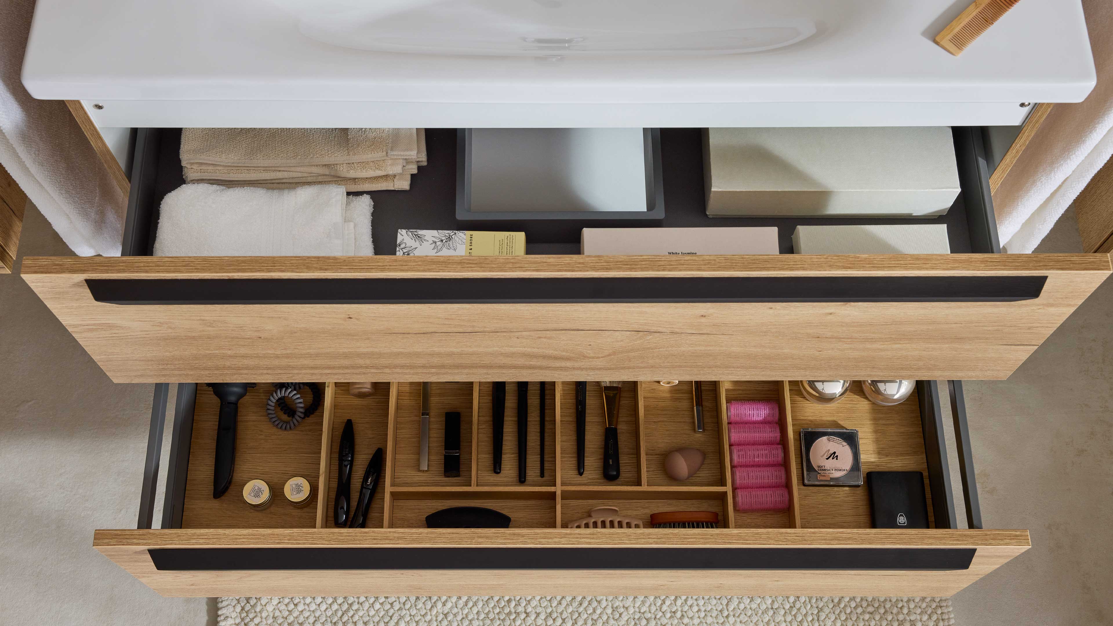 Geöffnete Eichen-Schubladen unter Waschbecken, obere mit Handtüchern und Kartons, untere mit Fächern für Pinsel, Bürsten und Kosmetik.