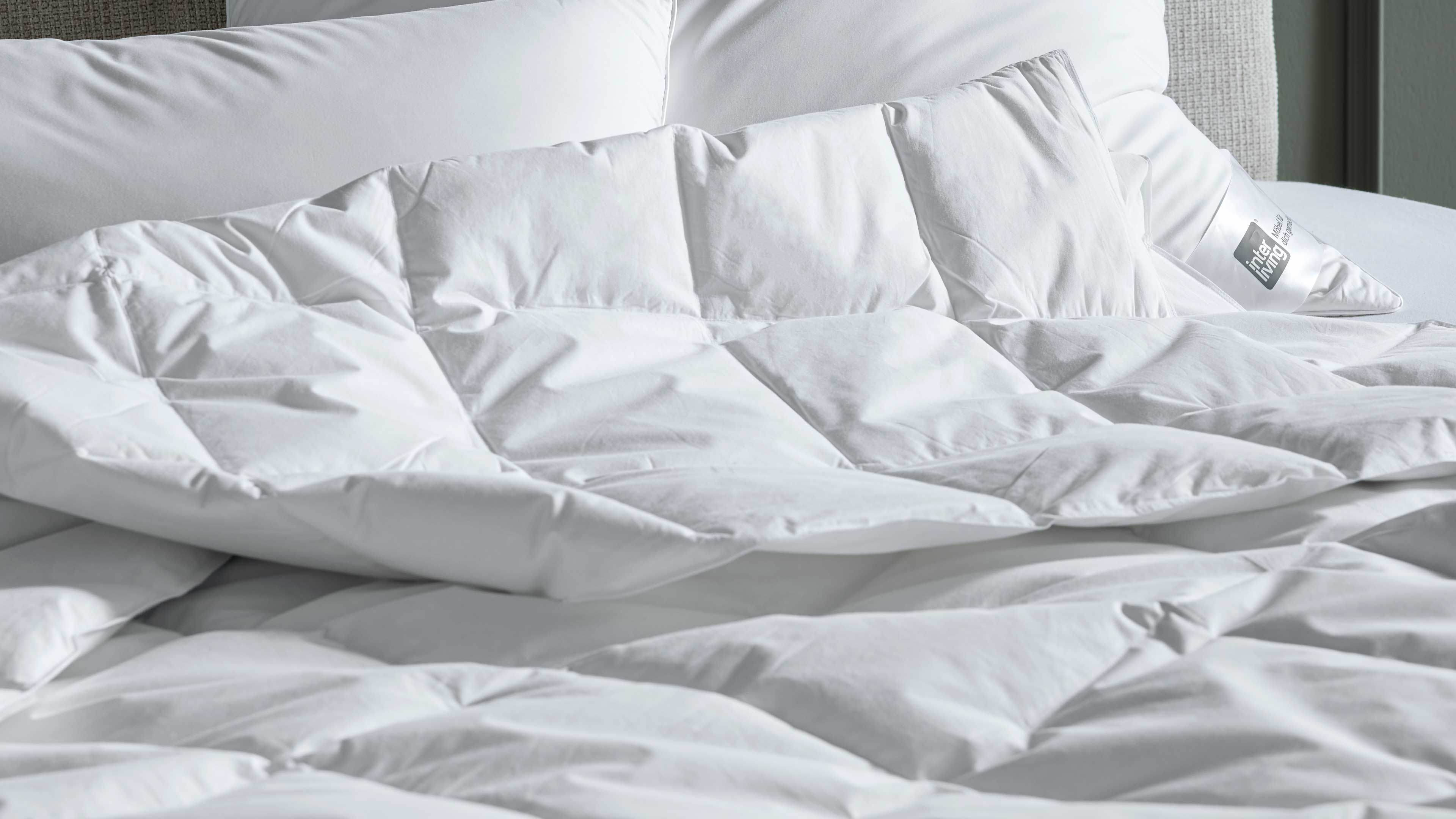 Weiße, gesteppte Bettdecke mit quadratischen Kammern locker aufgelegt, im Hintergrund mehrere weiße Kissen, rechts Kissen mit Etikett.