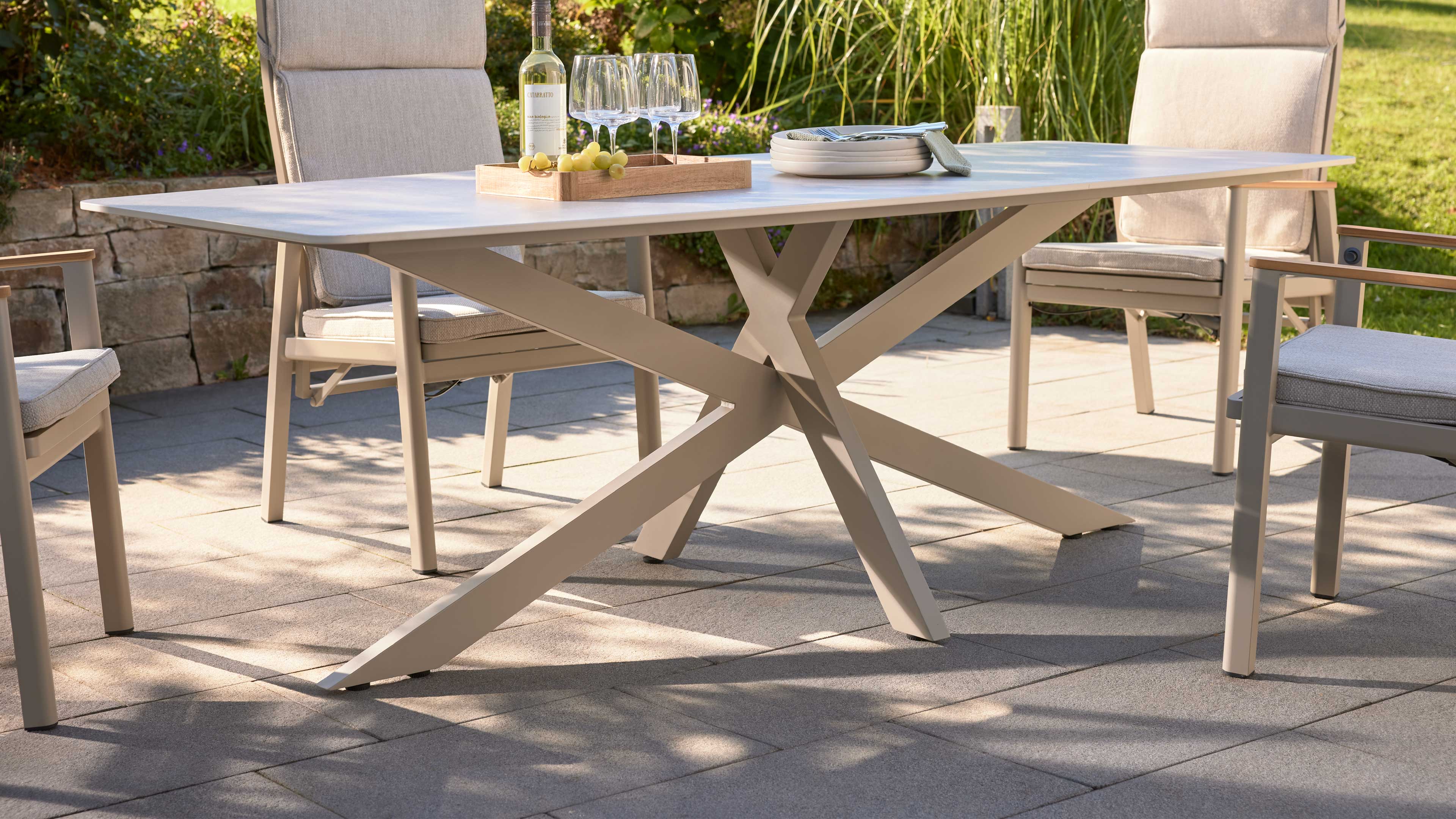 Beige Außen-Esstisch mit überkreuzten X‑Beinen auf gepflasterter Terrasse, dazu Polsterstühle und Tablett mit Weinflasche und Gläsern.