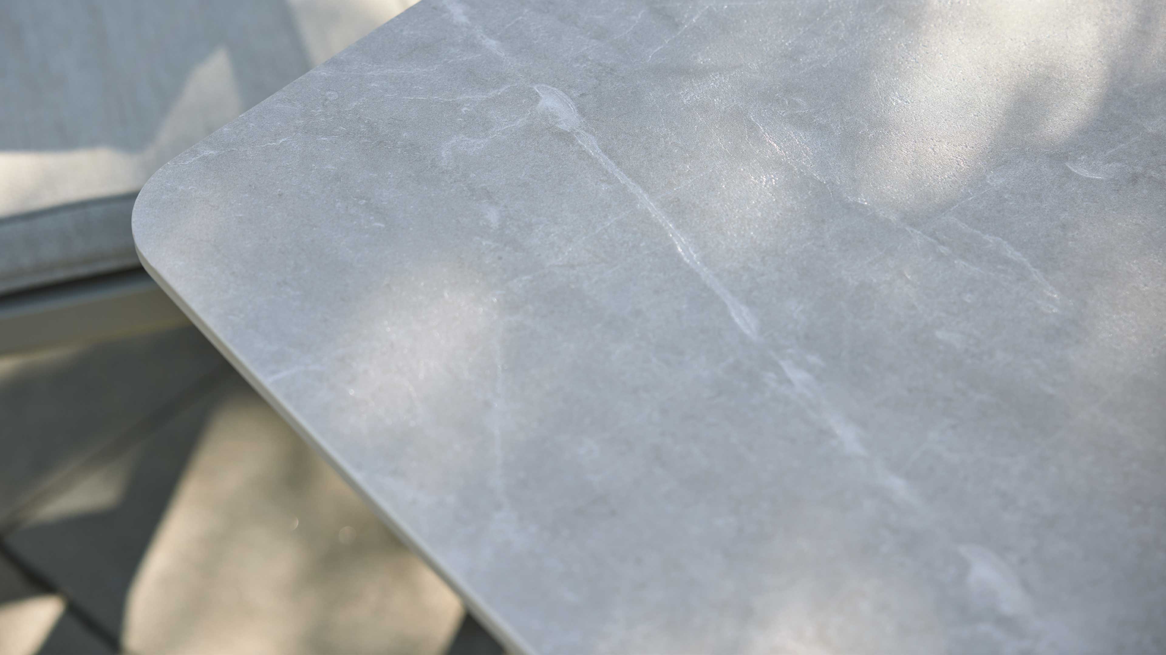 Graue Tischplatte mit feinen hellen Adern und abgerundeter Ecke, im Hintergrund ein helles Sitzkissen und Schatten.