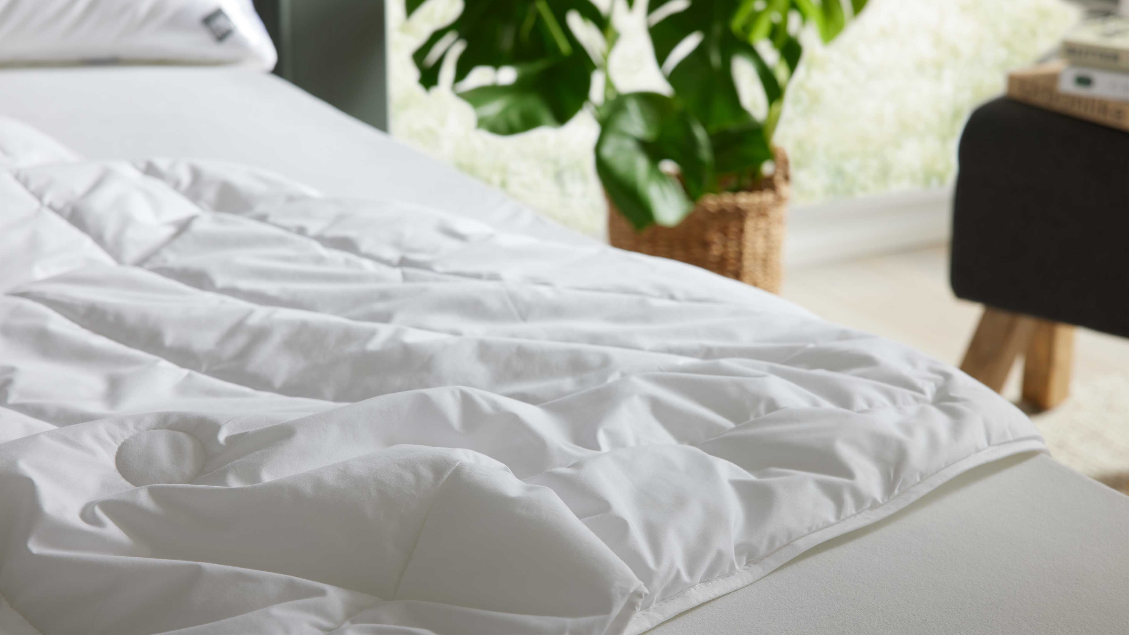 Weiße gesteppte Bettdecke auf Matratze, rechts ein dunkler Hocker mit Holzbeinen und im Hintergrund eine Grünpflanze im Korb.