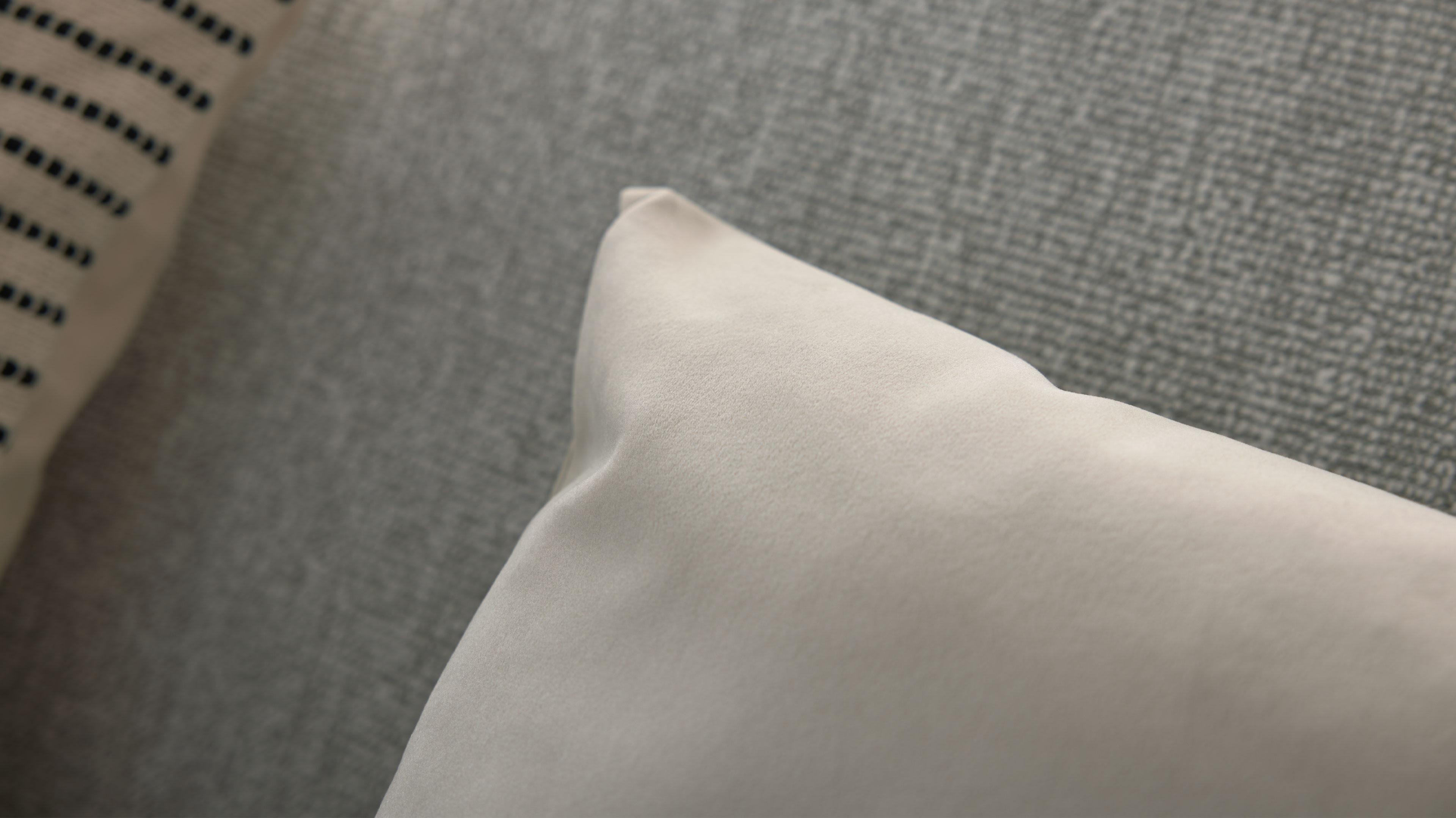 Hellbeiges Kissen im Vordergrund auf grauem, grob gewebtem Sofa; links ein zweites Kissen mit schwarzen Punkten.