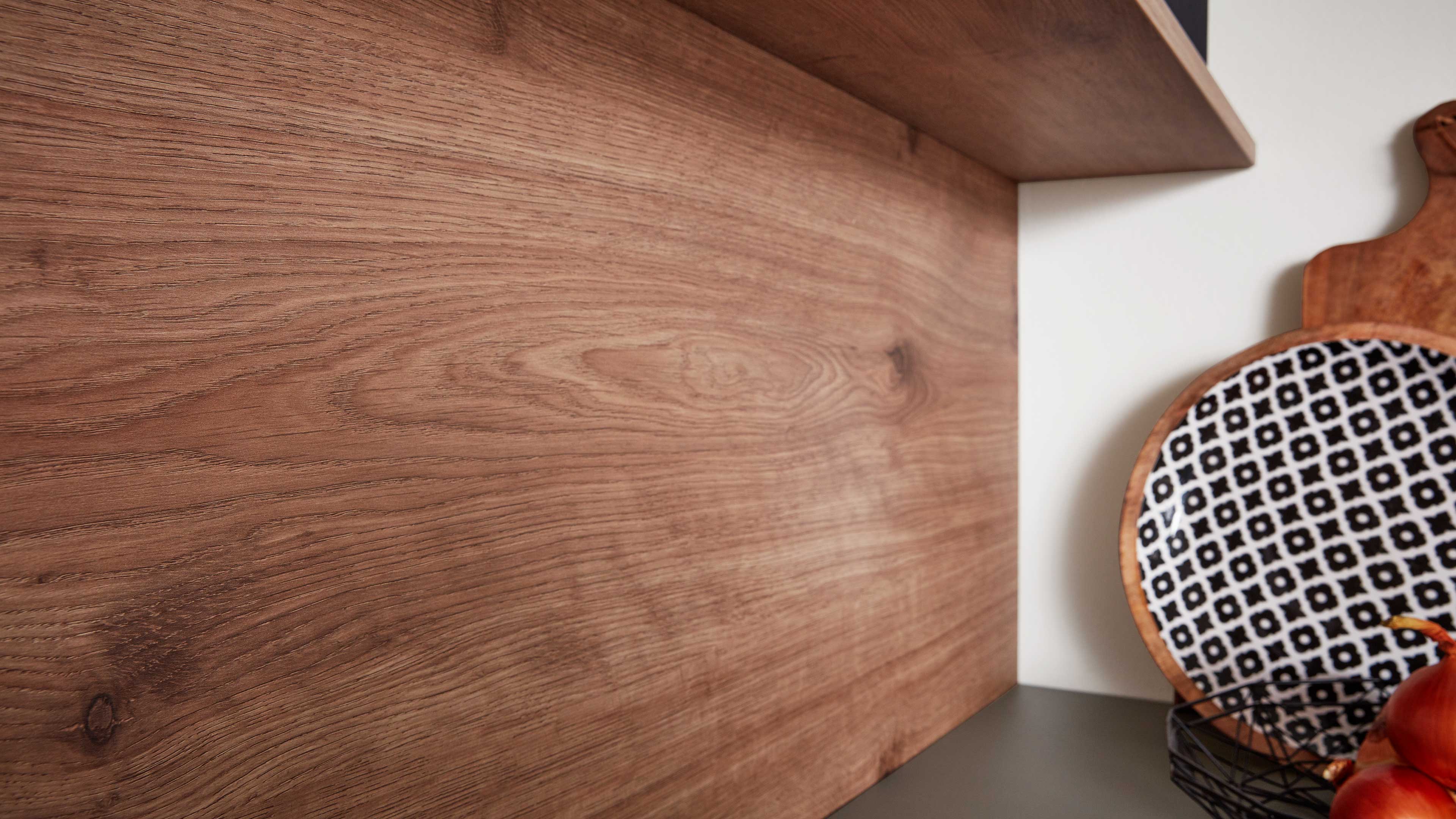 Holzrückwand mit sichtbarer Maserung, rechts rundes schwarz-weiß gemustertes Teller, Holzschneidebrett und Drahtkorb mit roten Zwiebeln.