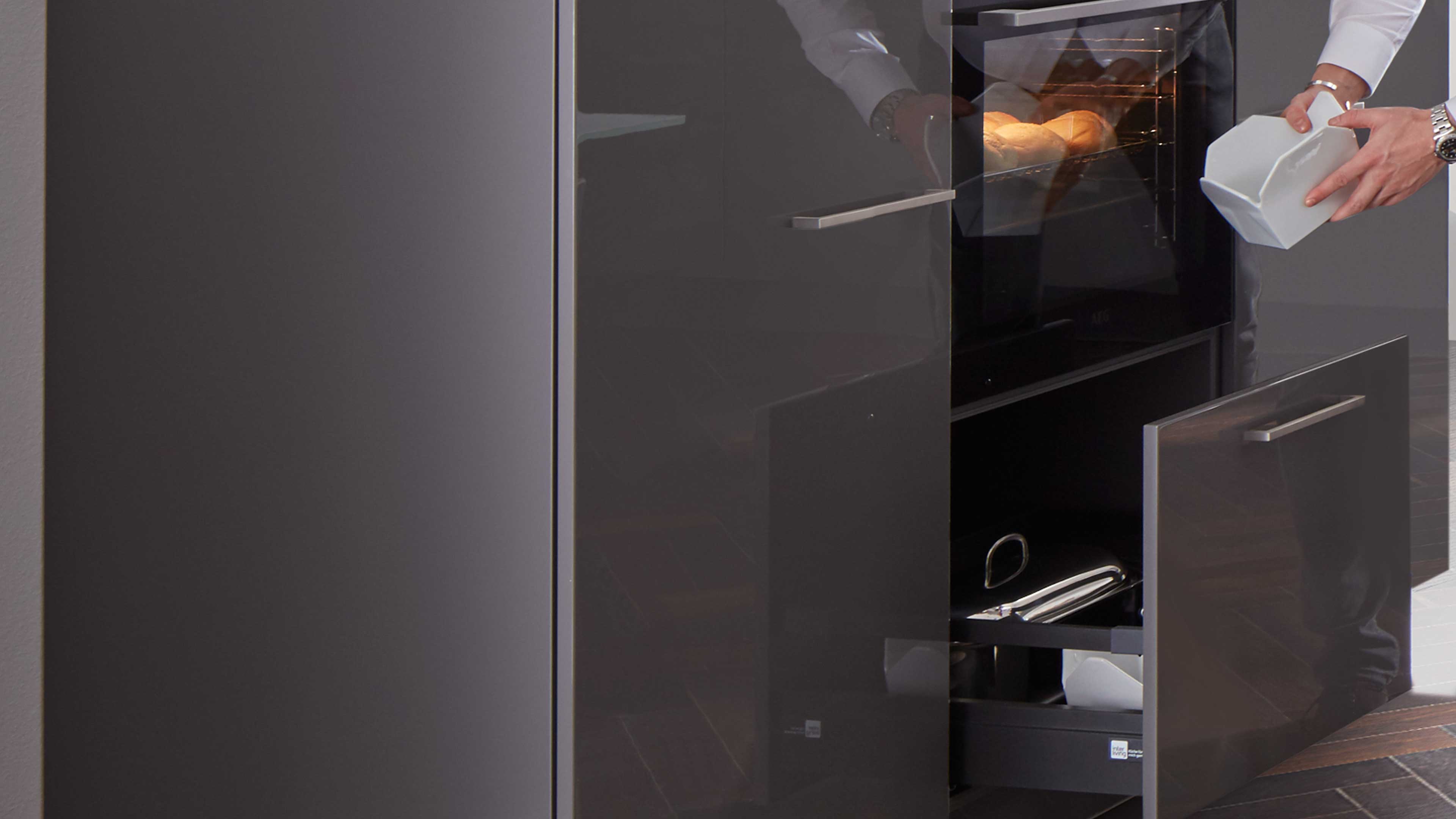 Küchenmöbel mit eingebautem Backofen, glänzende dunkelgraue Fronten, zwei schlanke Edelstahlgriffe und geöffnete Schublade mit Kochgeschirr.