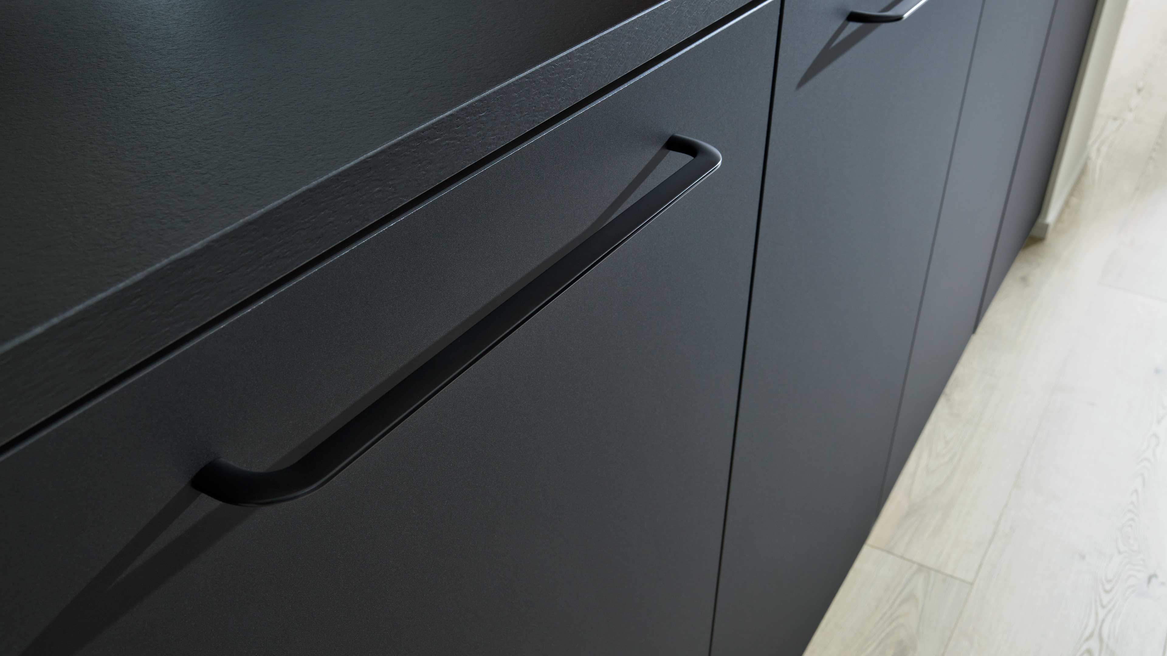 Dunkle Küchenfronten mit horizontal eingelassenem Griff und schmaler, strukturierter Arbeitsplattenkante, rechts heller Holzboden.