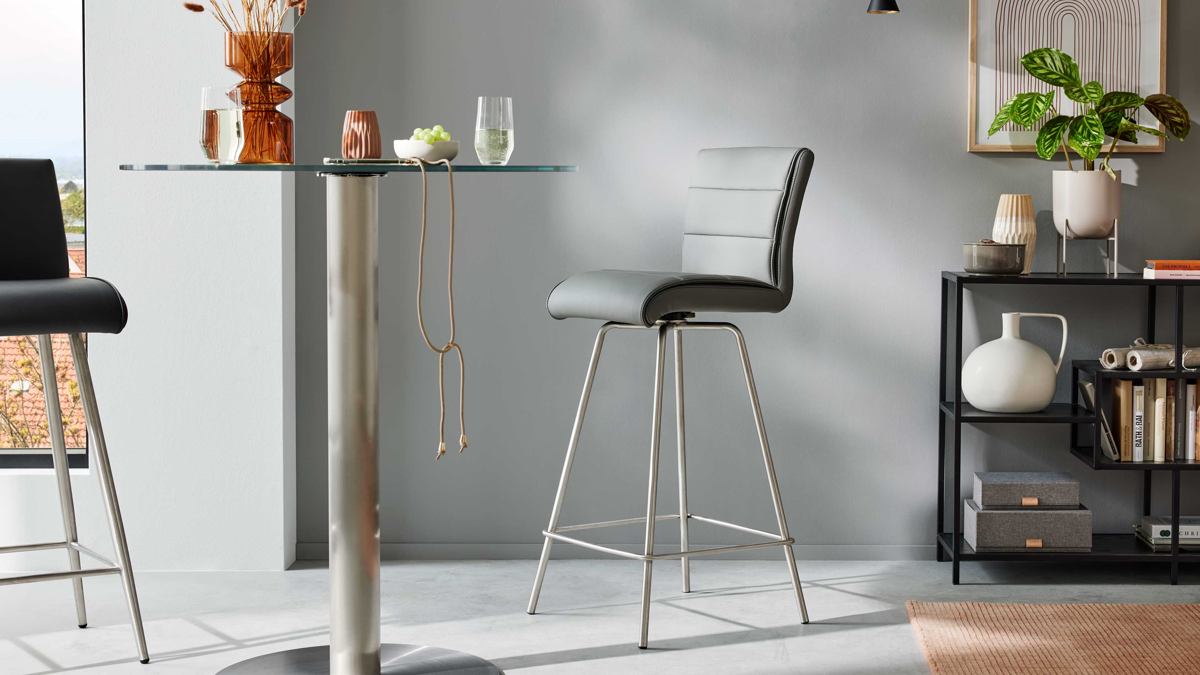Grauer Barhocker mit gepolstertem Sitz und Naht auf verchromten Beinen neben Glas-Stehtisch, rechts Regal mit Pflanze und Vasen.