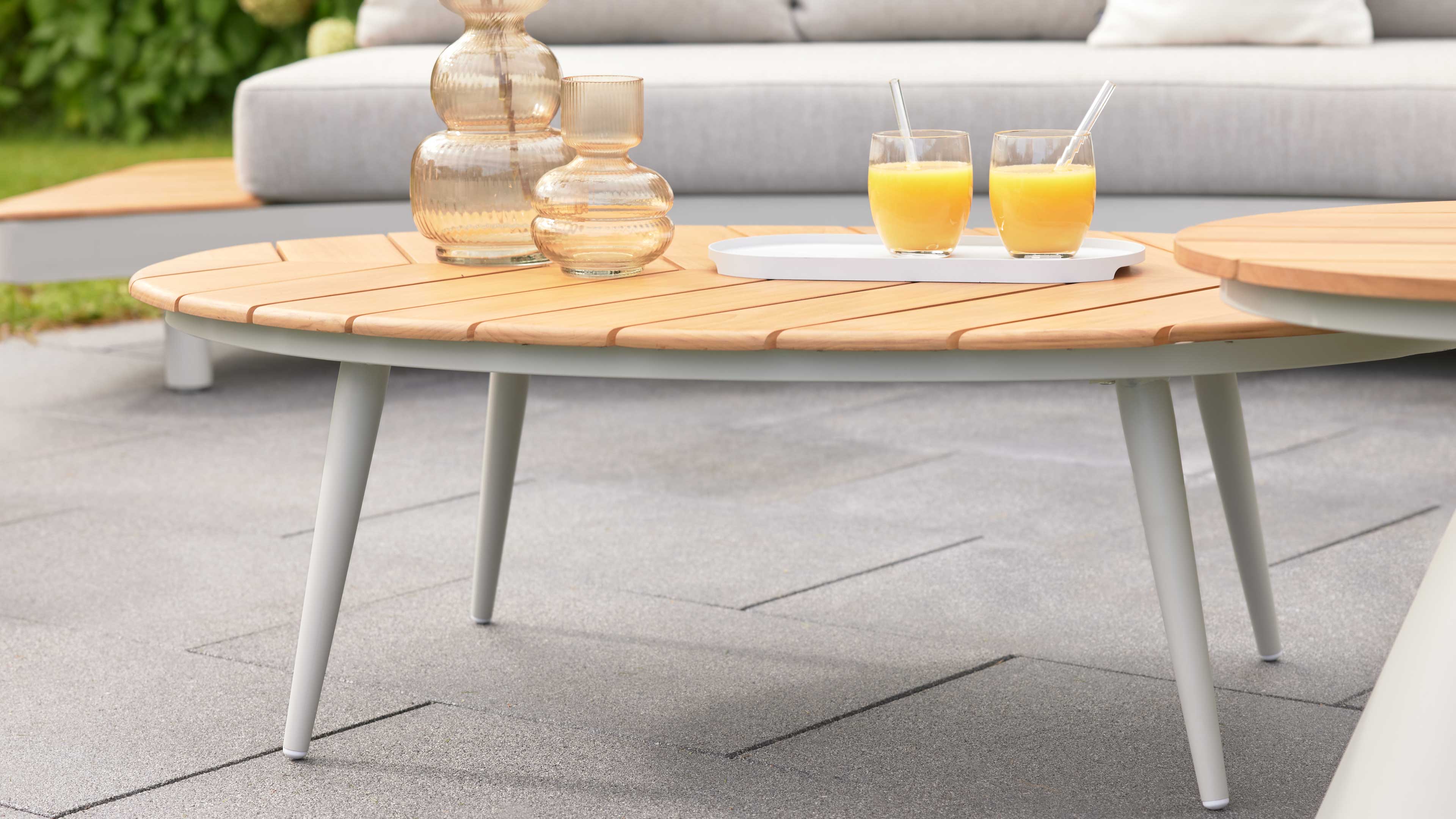 Runder Couchtisch mit hellem Holzbelag und weißen Metallbeinen, darauf zwei gerippte Glasvasen und ein Tablett mit zwei Orangensaftgläsern.