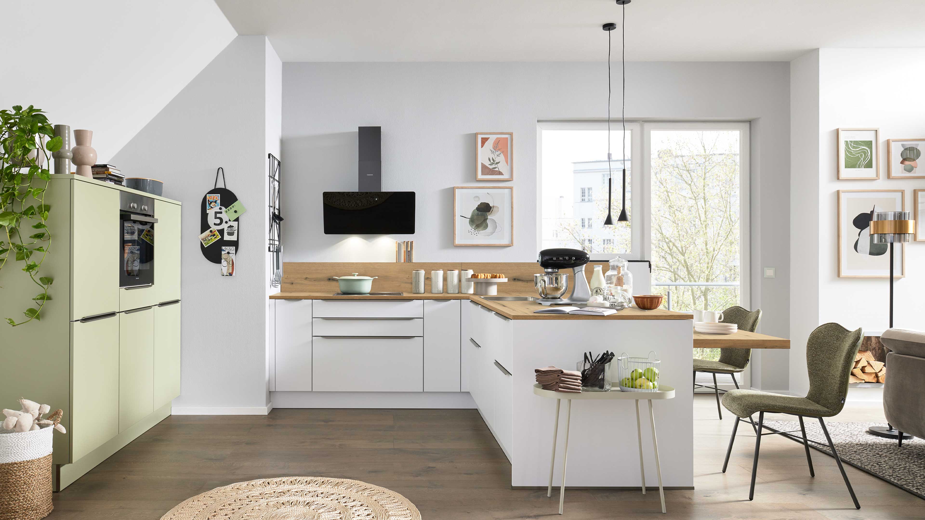Weiße L‑Küche mit Holzarbeitsplatte und schwarzer Dunstabzugshaube, grünem Hochschrank, Küchenmaschine und Essplatz mit grünen Stühlen.