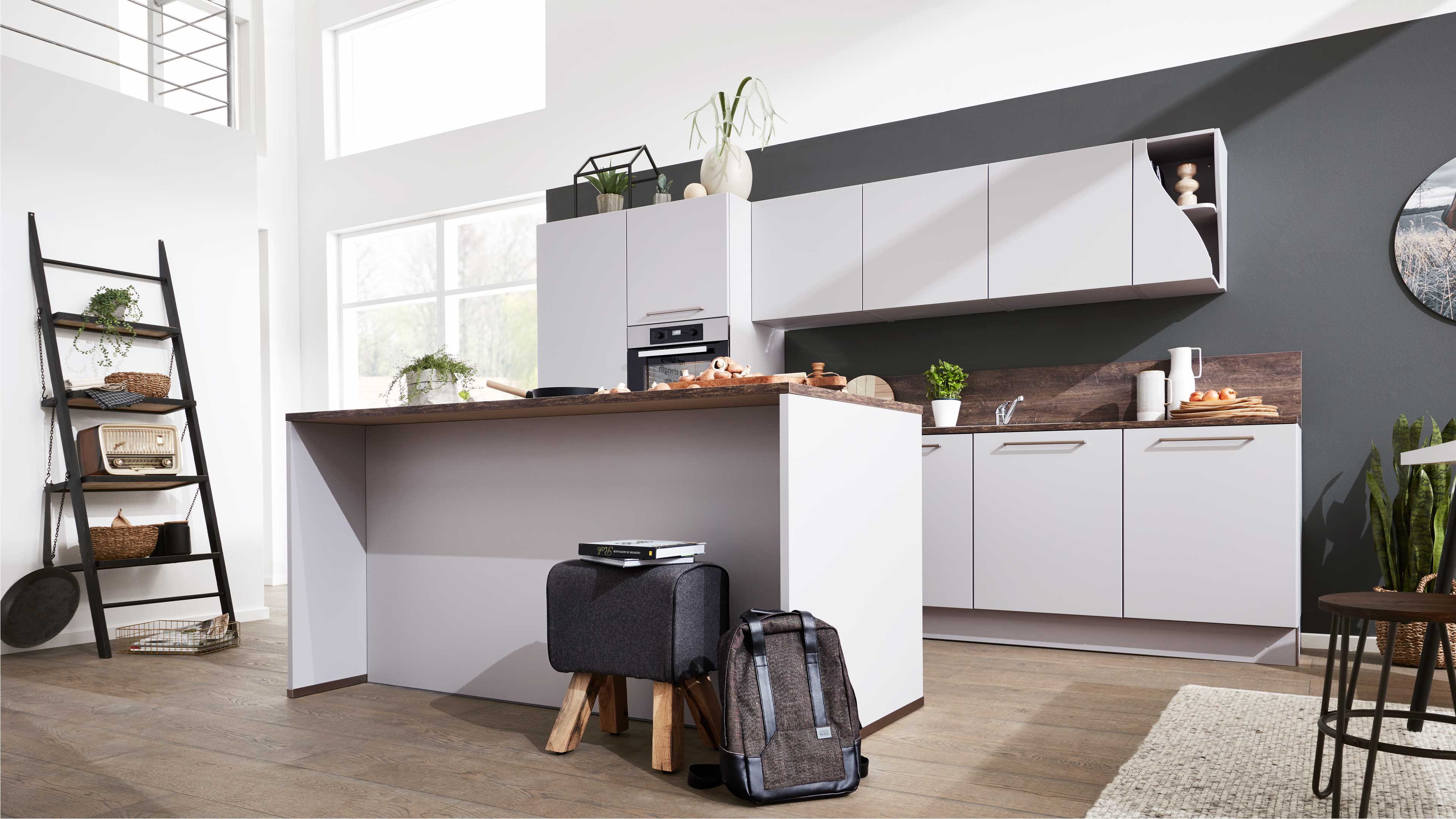 Weiße Kücheninsel mit Holzarbeitsplatte, davor grauer Hocker und Rucksack, im Hintergrund Oberschränke und Leiterregal.