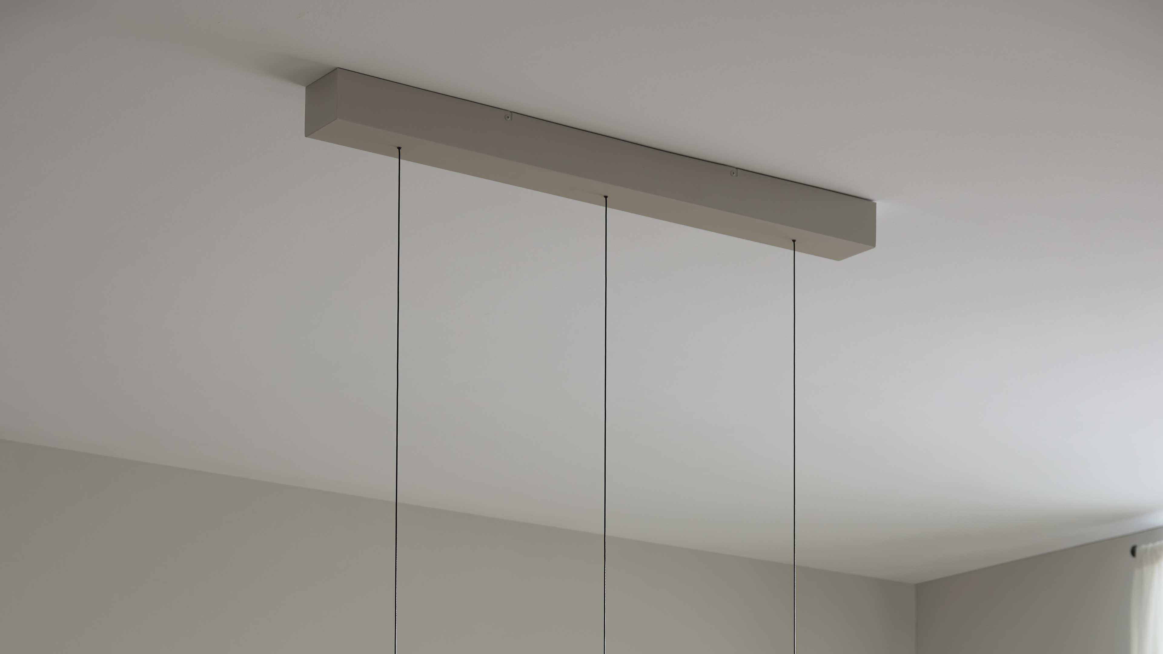 Rechteckige Deckenleiste mit drei dünnen schwarzen Abhängungen, darunter helle Zimmerwand und sichtbare Fensterkante.